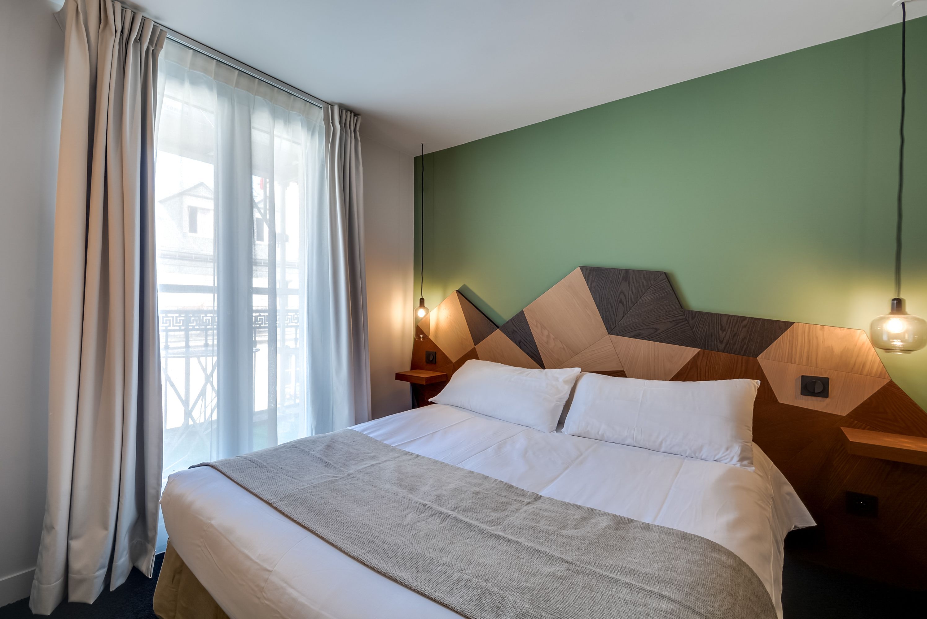 A hotel room with a bed, curtains, a nightstand, and a wall-mounted lamp.