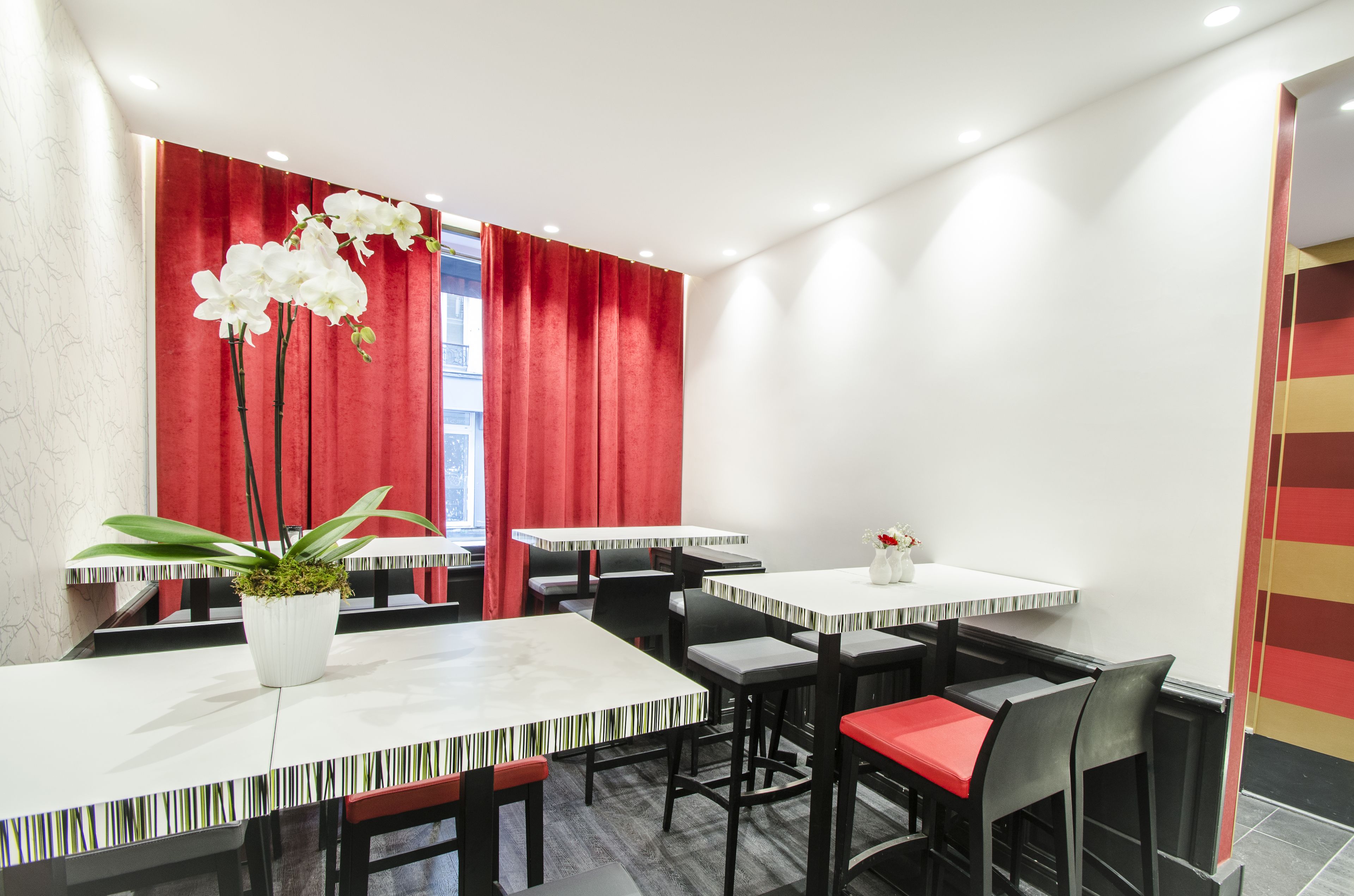 A modern dining area with white tables and red curtains, featuring a potted plant and a vase with flowers.