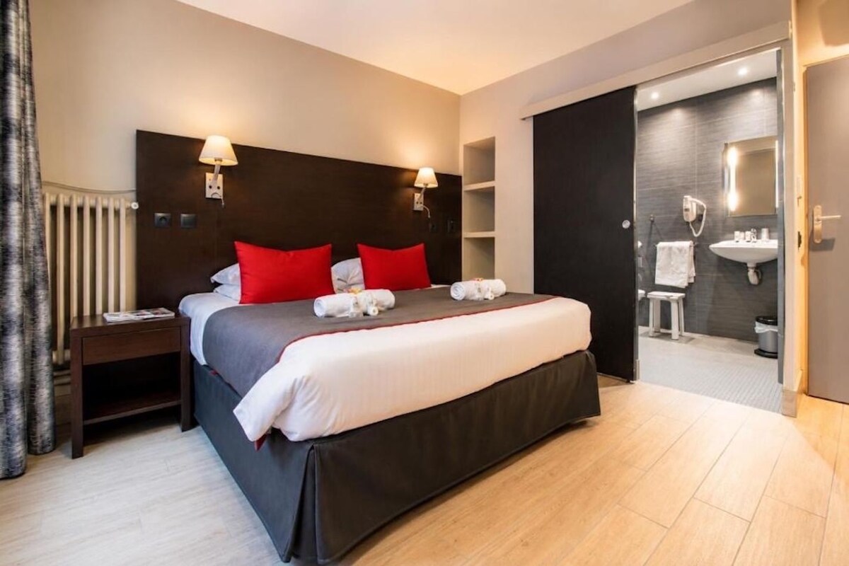 A hotel room with a large bed, bedside lamps, a nightstand, a bathroom with a sink and mirror, and a door leading to another room.