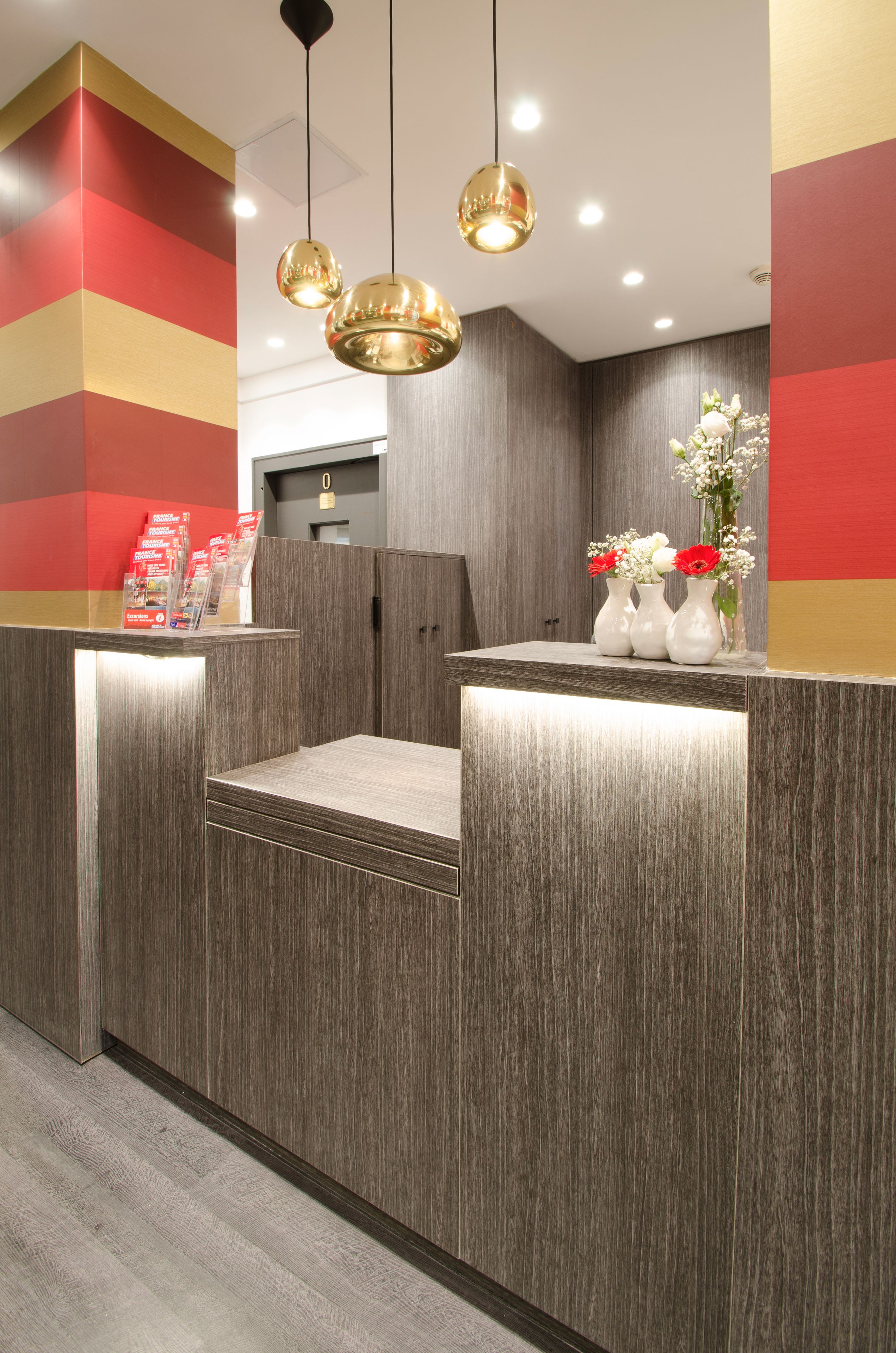 A reception desk with a modern design, featuring a wooden finish and a grey cushioned seating area. There are three pendant lights hanging above the desk, and a vase with flowers on the right side.