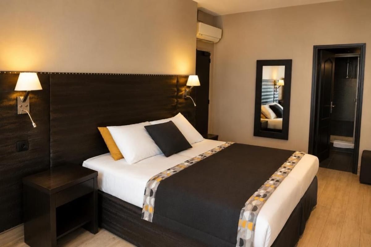 A hotel room with a large bed, bedside lamps, a mirror, and an air conditioning unit.