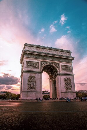 A large stone arch monument with intricate carvings and statues, surrounded by a paved area with people and a scooter. - Hotel Migny Opera Montmartre (Paris)
