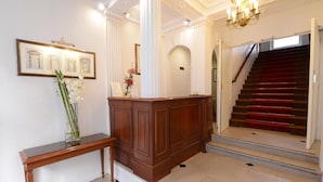A reception area with a wooden desk, a bench, a vase with flowers, framed artwork, and a staircase with red carpet leading to an upper floor. - Hotel Impérial Paris (Paris)