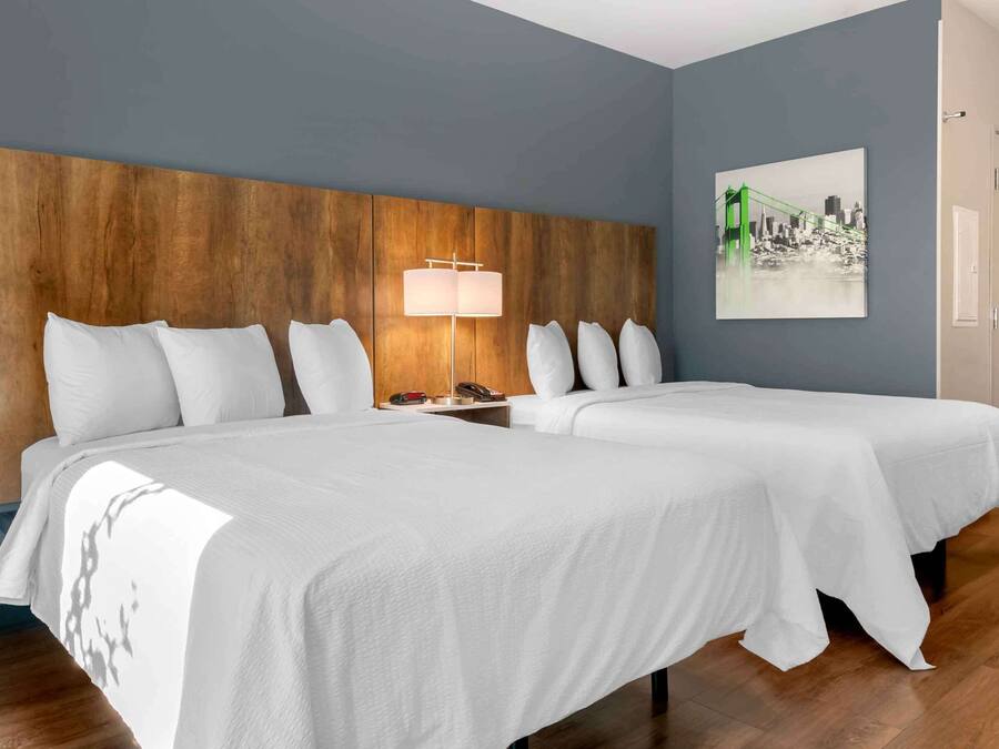 Habitación de hotel con dos camas, cabecero de madera, mesita de noche con lámpara y un cuadro de paisaje urbano en la pared.