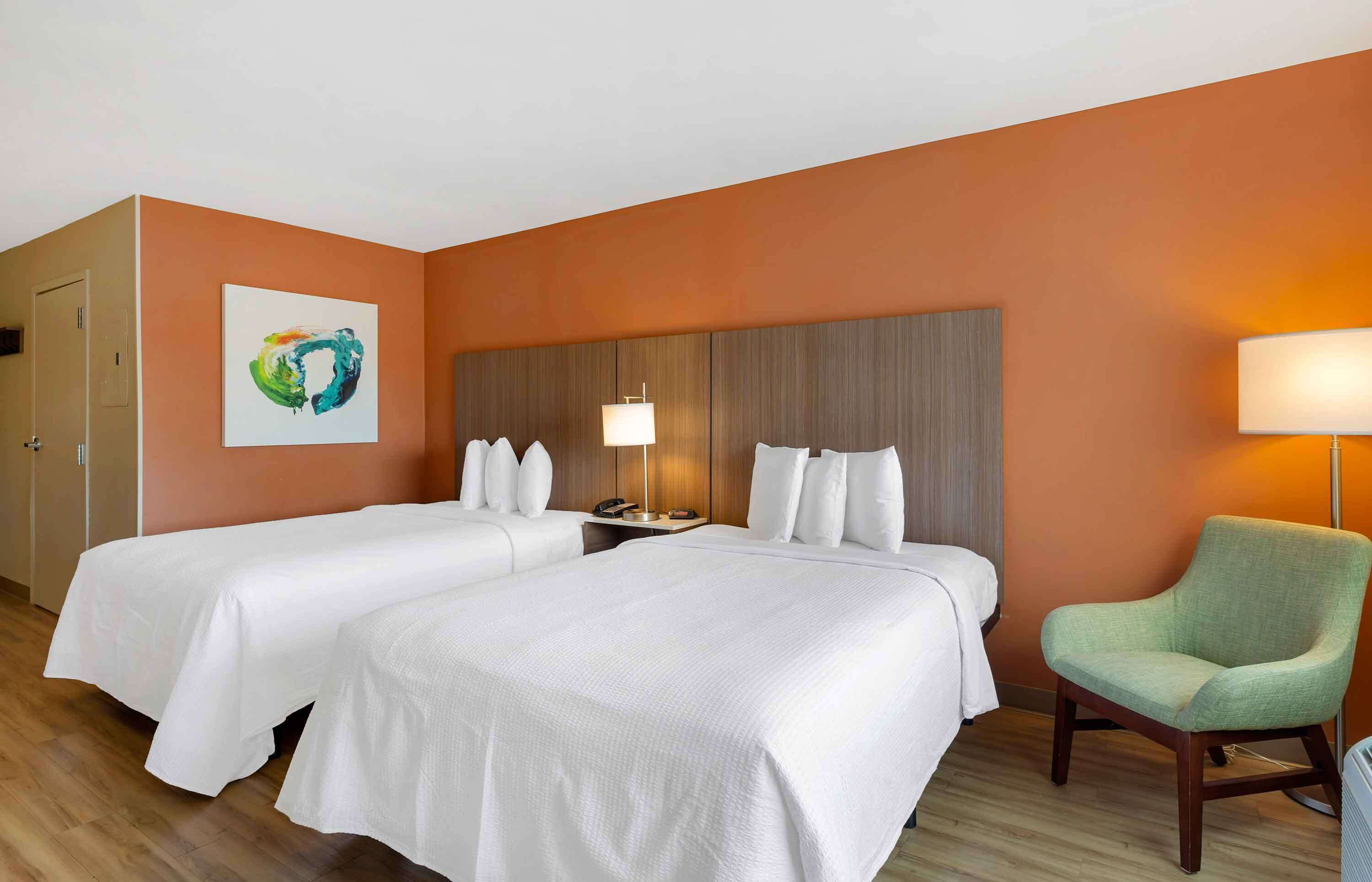 Una habitación de hotel con dos camas, un sillón verde, una mesita de noche con una lámpara y una obra de arte en la pared.