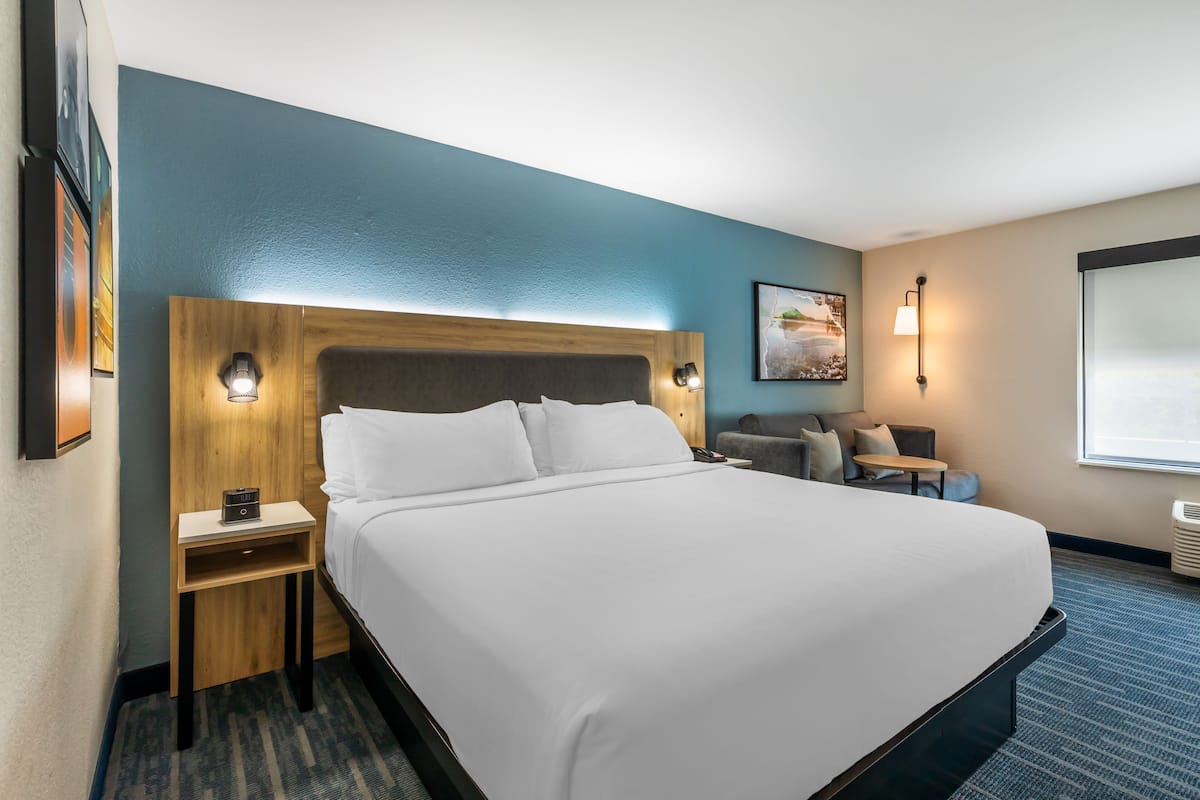 A hotel room with a large bed, a wooden headboard, a nightstand, a small table, a chair, and a window with a view.