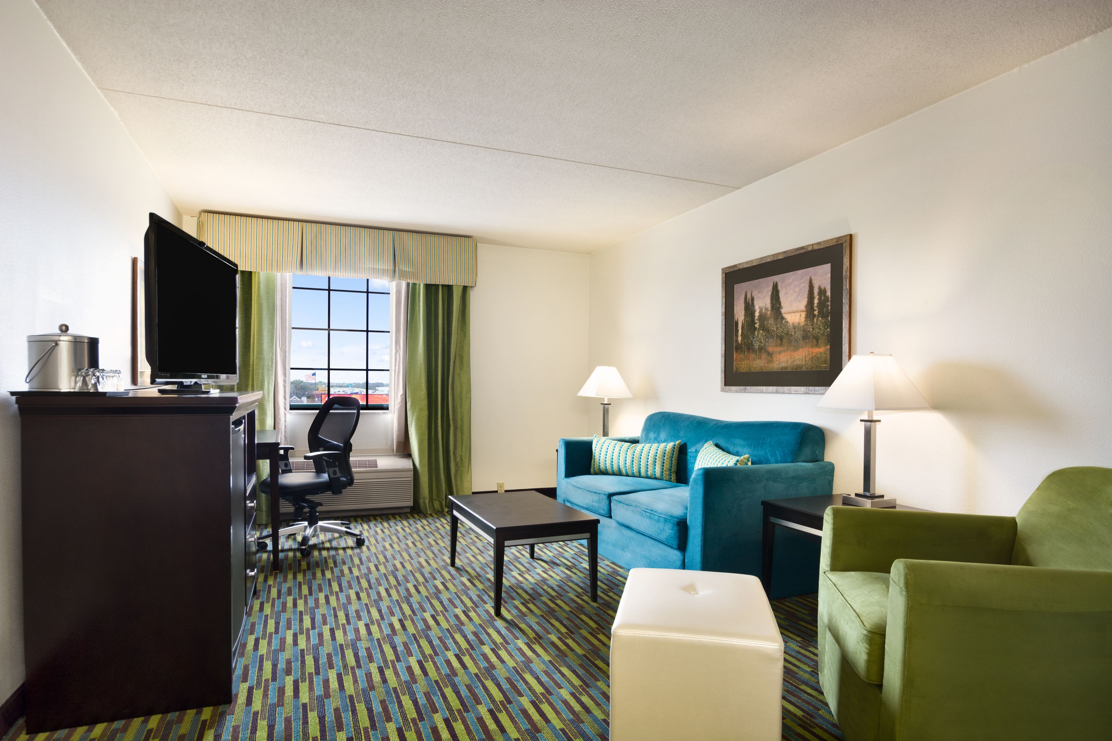 A hotel room with a desk, a blue sofa, a green armchair, a white ottoman, and a television.