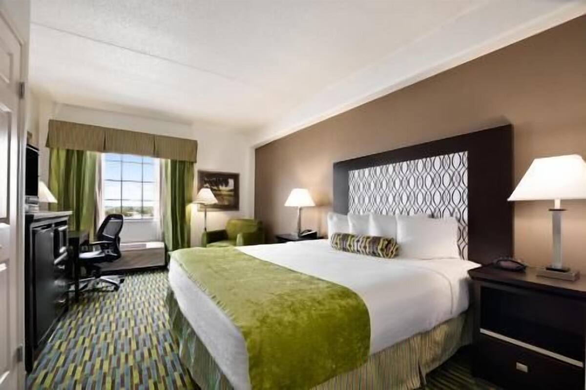 A hotel room with a large bed, two bedside tables, a desk with a chair, a window with curtains, and a patterned carpet.