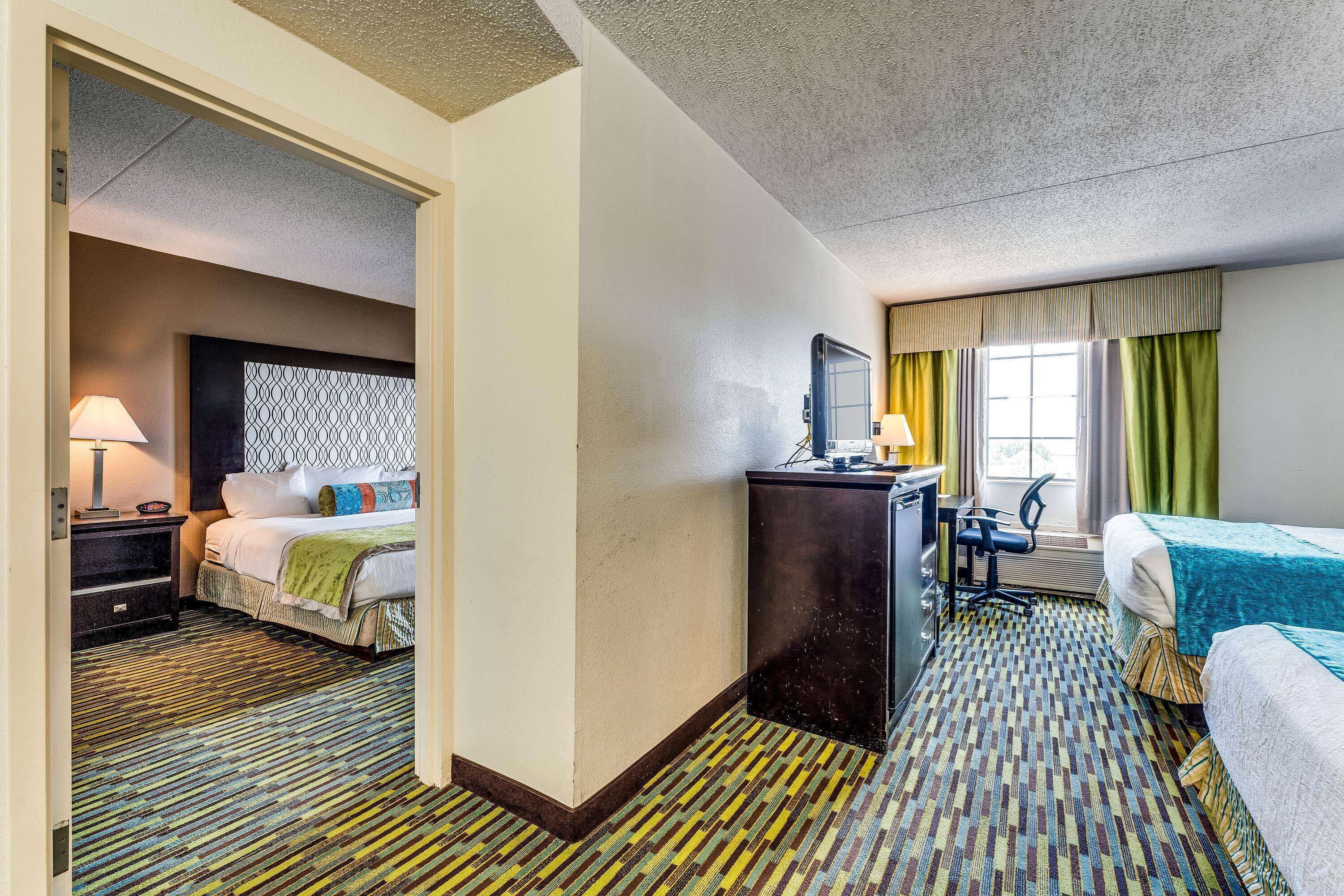 A hotel room with a bed, desk, and chair, a patterned carpet, and a window with curtains.