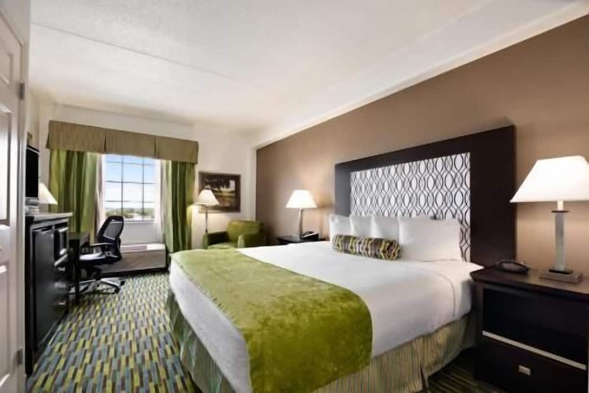 A hotel room with a large bed, two bedside tables, a desk with a chair, a window with curtains, and a patterned carpet.