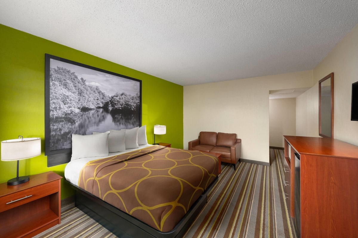 A hotel room with a large bed, a green accent wall, a framed picture, a desk with a lamp, a brown leather chair, and a wooden dresser.