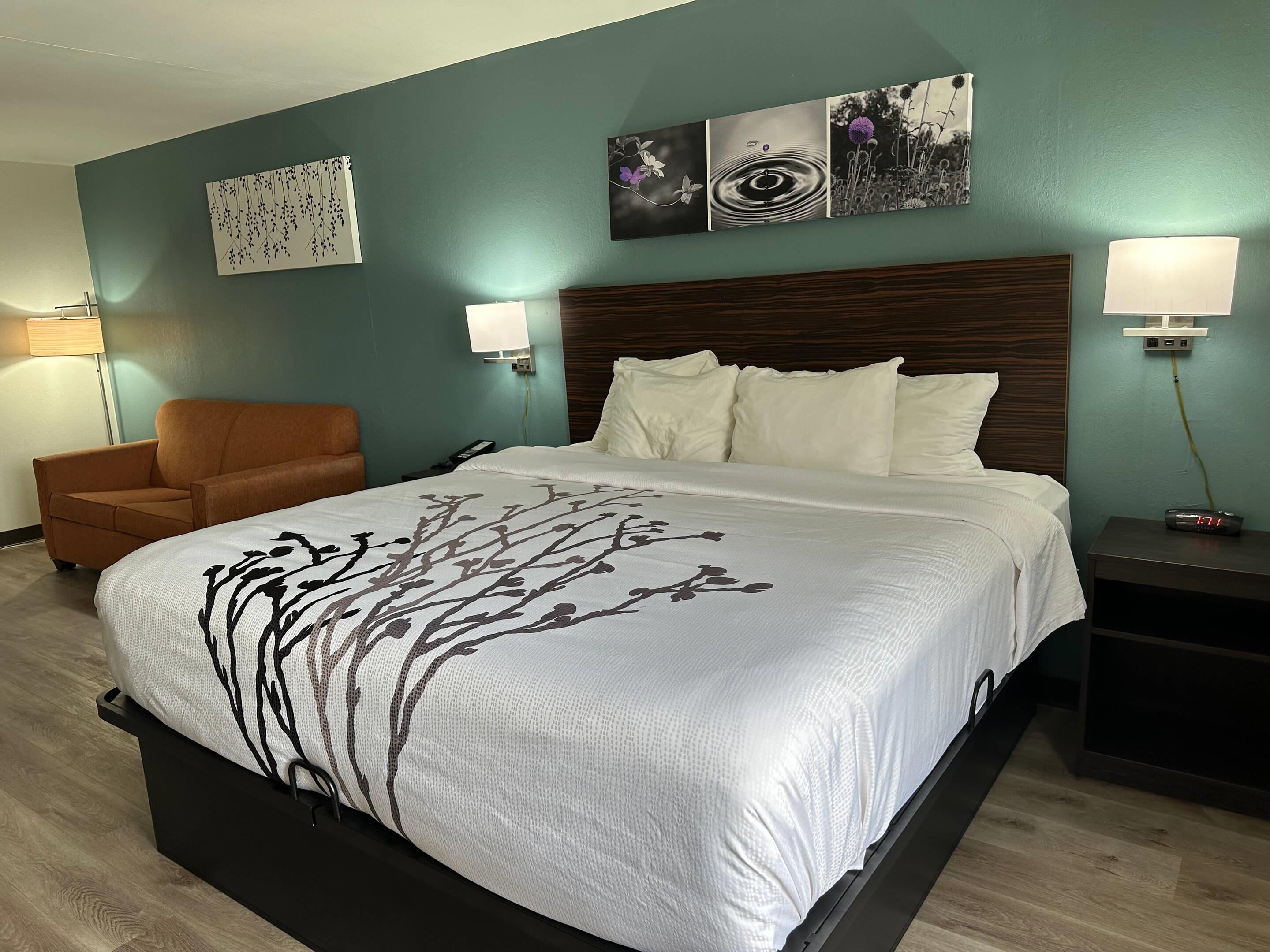 A hotel room with a large bed, a chair, a nightstand, and wall art.
