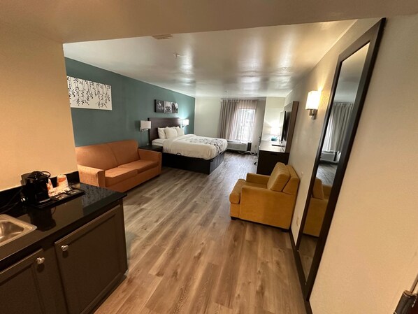 A hotel room with a bed, a sofa, a chair, a desk with a coffee maker, and a mirror.