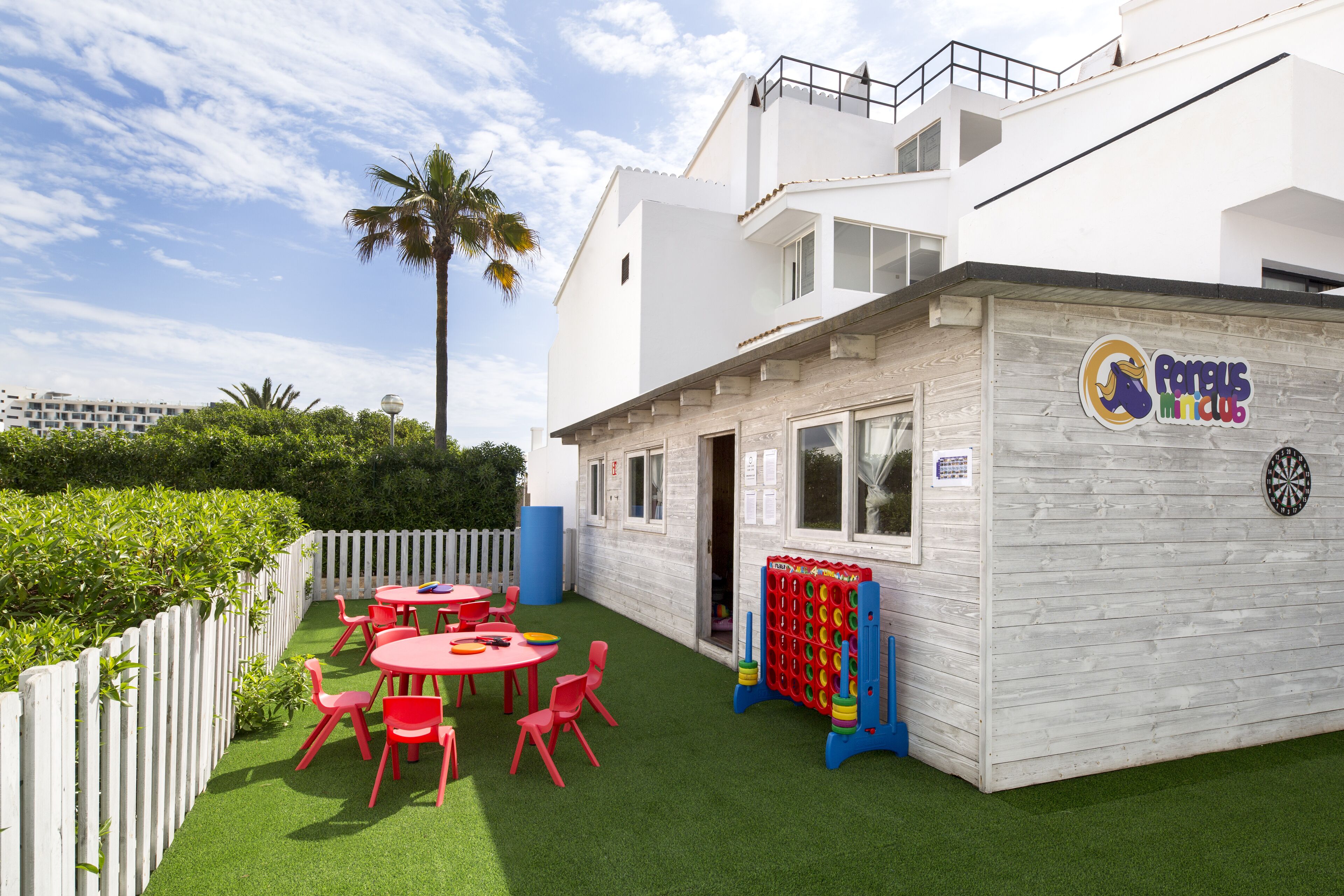 A children's play area with red tables and chairs, a dartboard, and a white building with a sign reading "Famous Club".