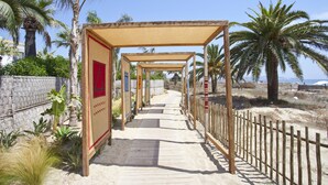 Un paseo marítimo de madera con zonas sombreadas, palmeras y una playa arenosa.