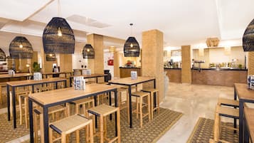 Um restaurante com mesas e cadeiras de madeira, luminárias negras pendentes e piso em padrão herringbone.