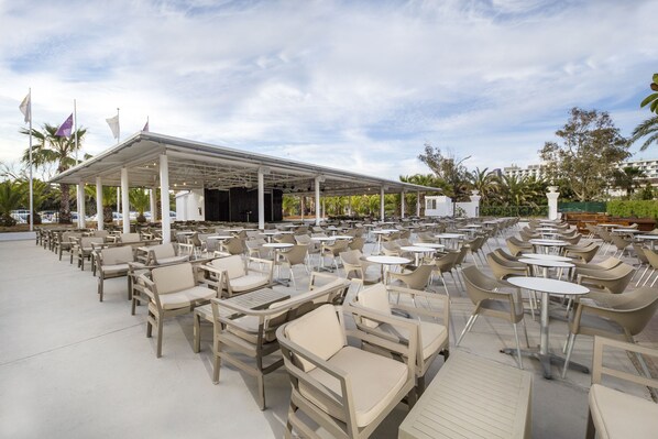 Un espace extérieur aménagé avec des tables et des chaises, protégé par un auvent, et entouré de palmiers, avec un ciel dégagé.