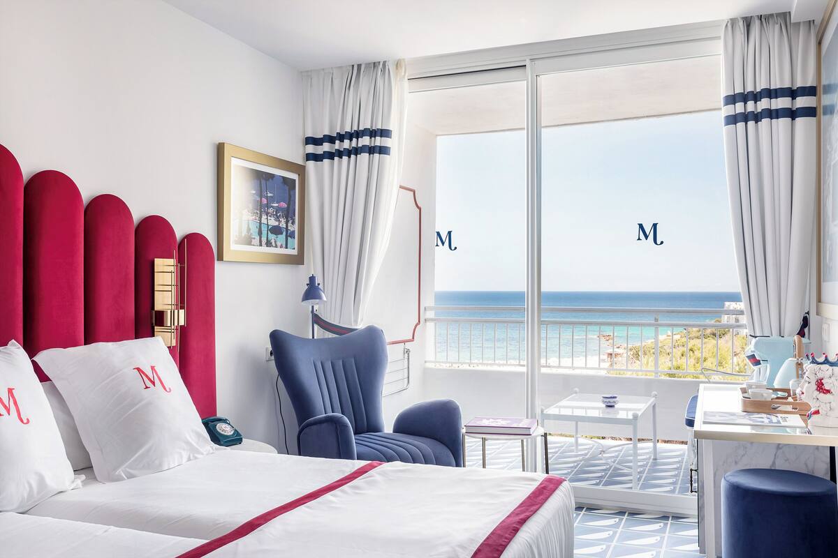 A hotel room with a bed, a chair, a table, and a view of the sea.