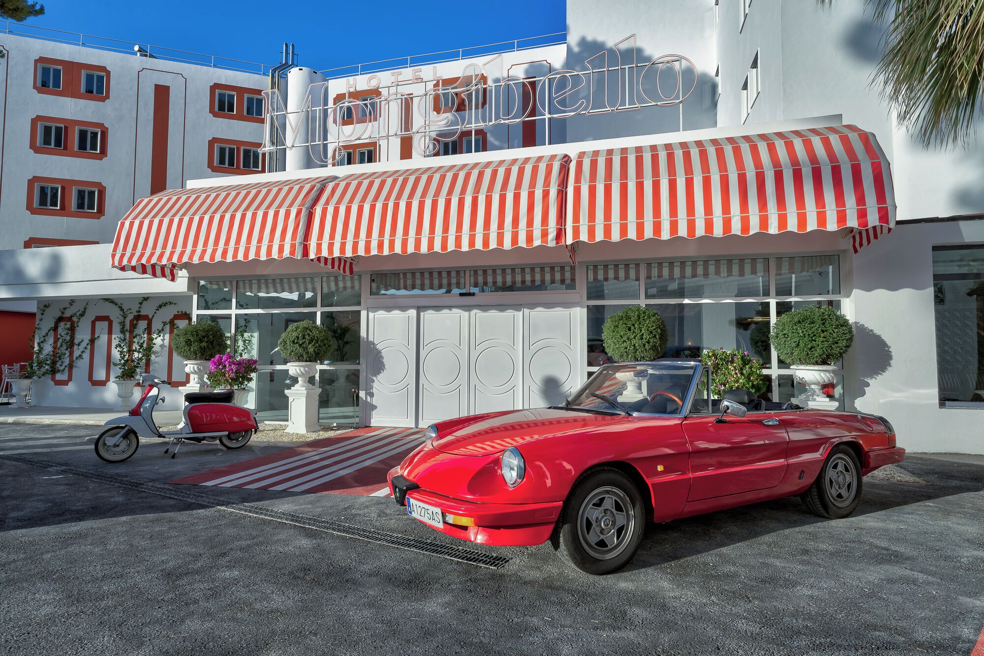 Een rode cabrio geparkeerd voor een hotel met een gestreept luifel en een bord waarop 