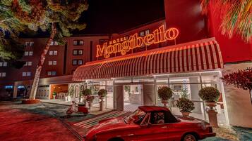 A hotel with a neon sign, a red car, and potted plants in front.