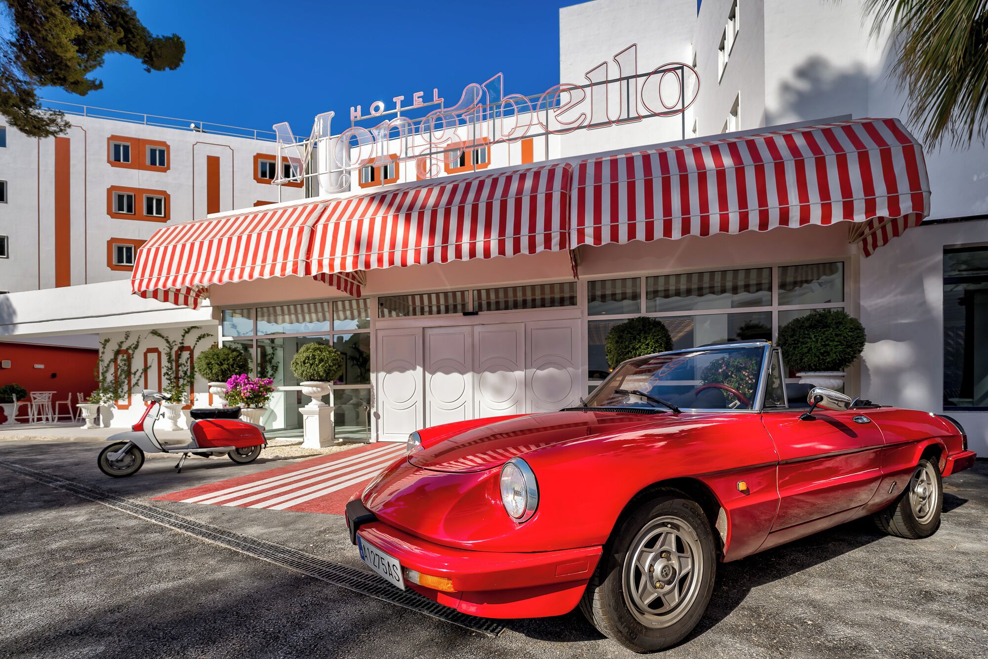 A red convertible parked in front of a hotel with a striped awning and a sign that reads "HOTEL MONTEBELLO".