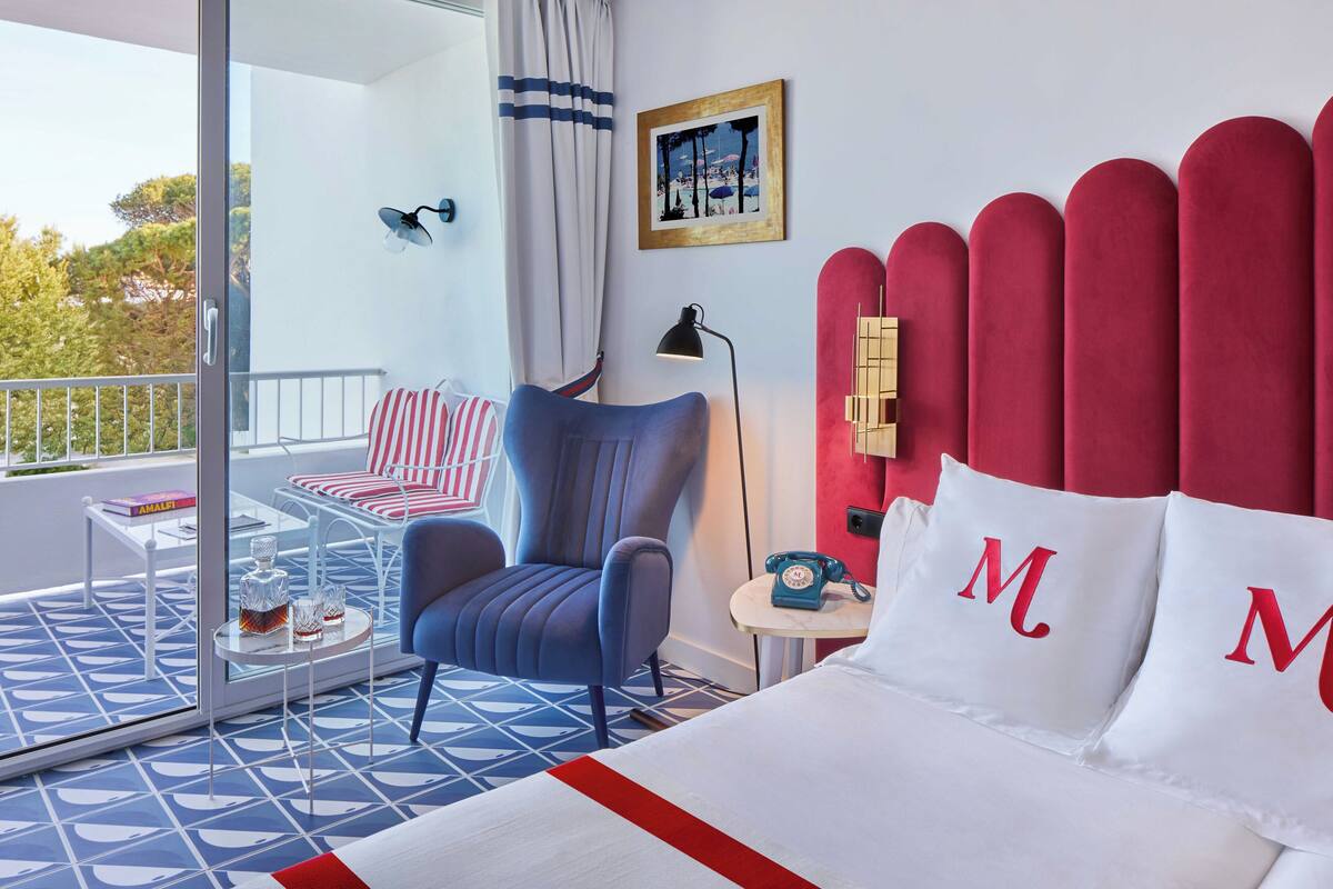 A hotel room with a bed, a chair, a small table, a phone, and a window with a view of trees.