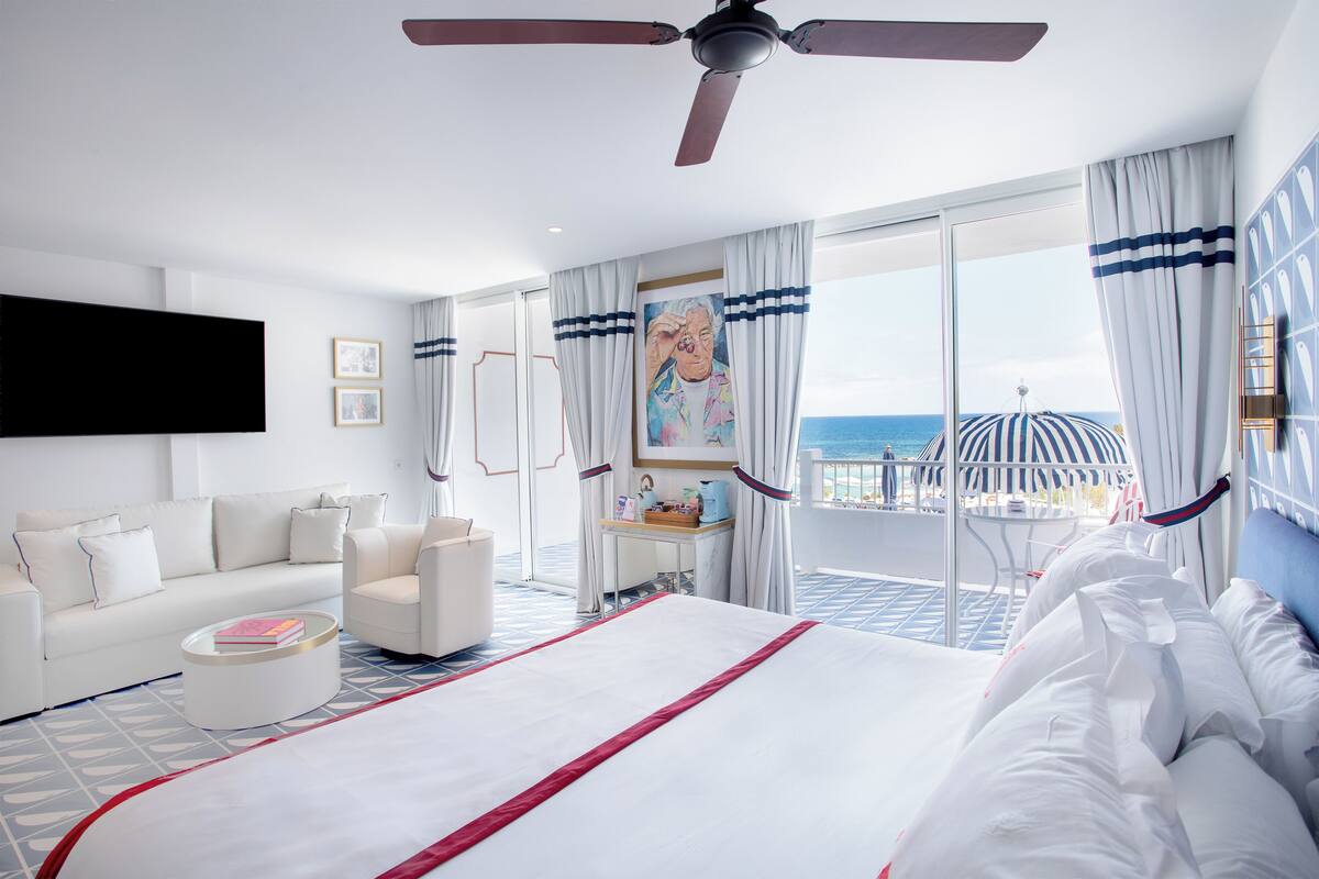A modern hotel room with a large bed, a sofa, a coffee table, and a TV. The room has a balcony with a view of the sea.