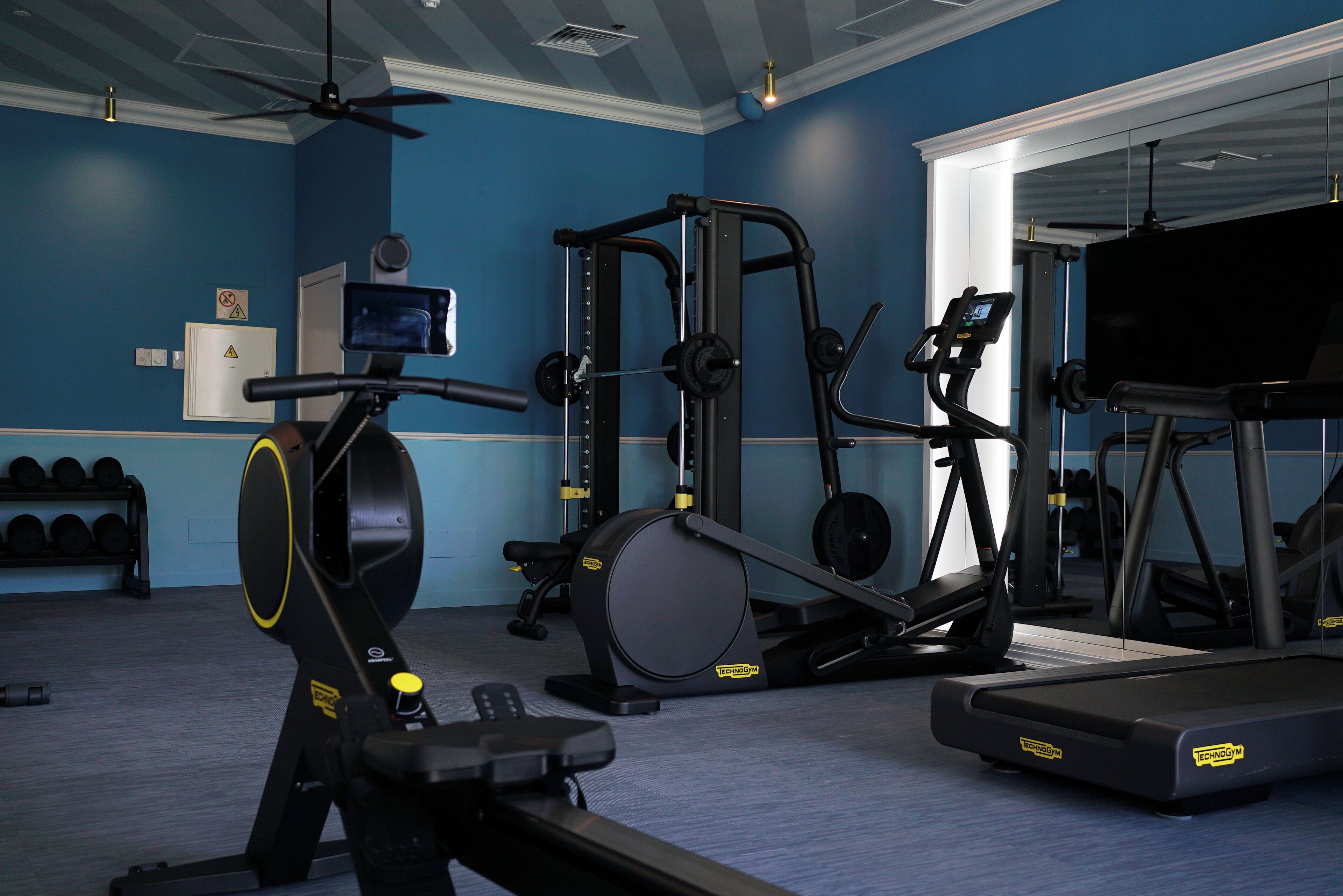 Ein Fitnessraum mit Trainingsgeräten, Laufband und Wandfernseher.