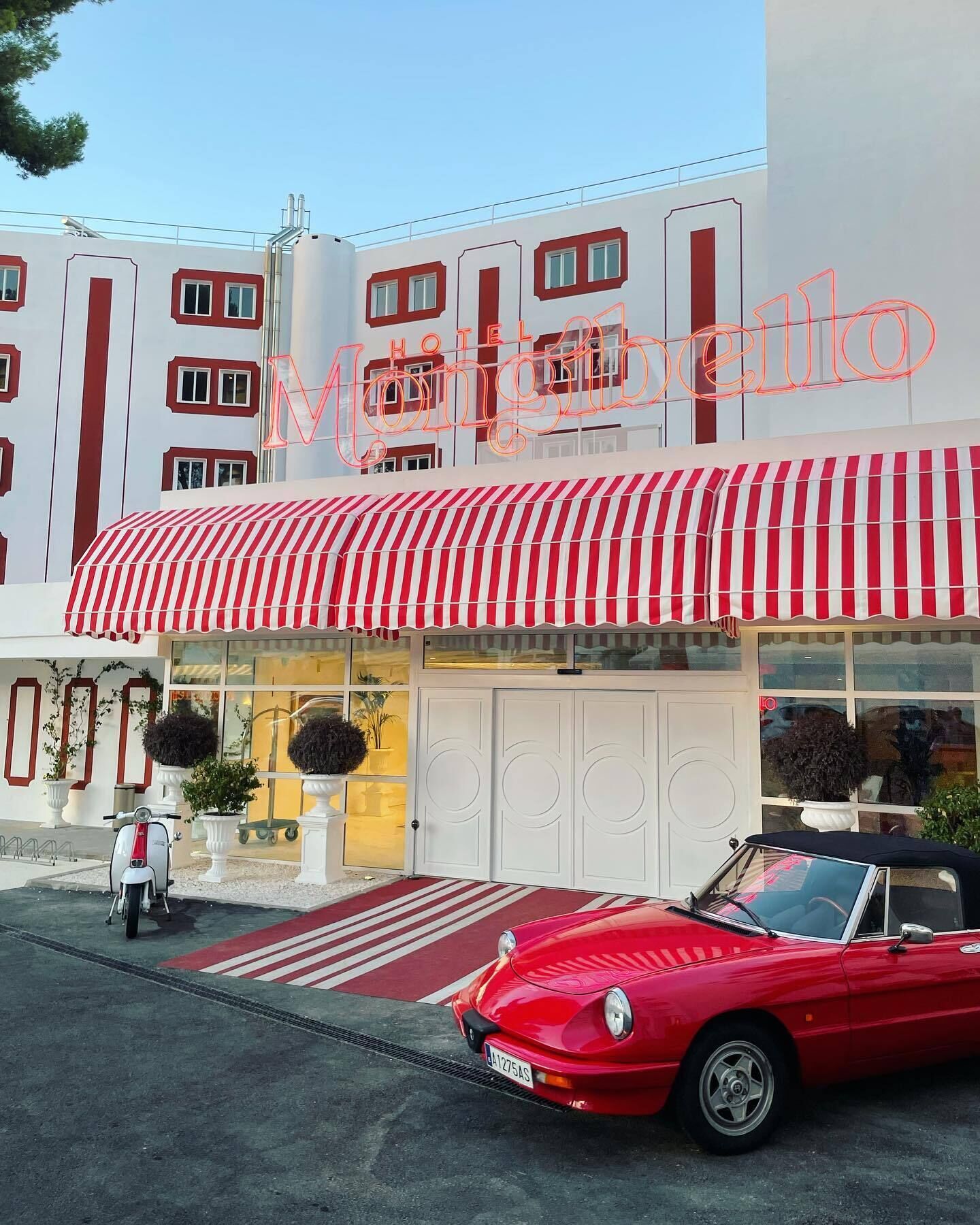 A hotel with a red and white striped awning, a classic red convertible parked in front, and a modern building facade with a neon sign.