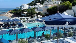 Ein Poolbereich mit Liegestühlen und Sonnenschirmen mit Blick auf einen Strand und Gebäude an einem Hügel.