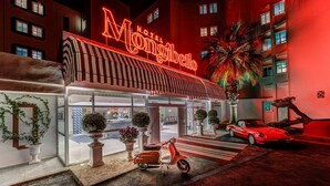 Ein Hotel Eingang mit einer Neonreklame, einer Palme und einem klassischen roten Auto davor.