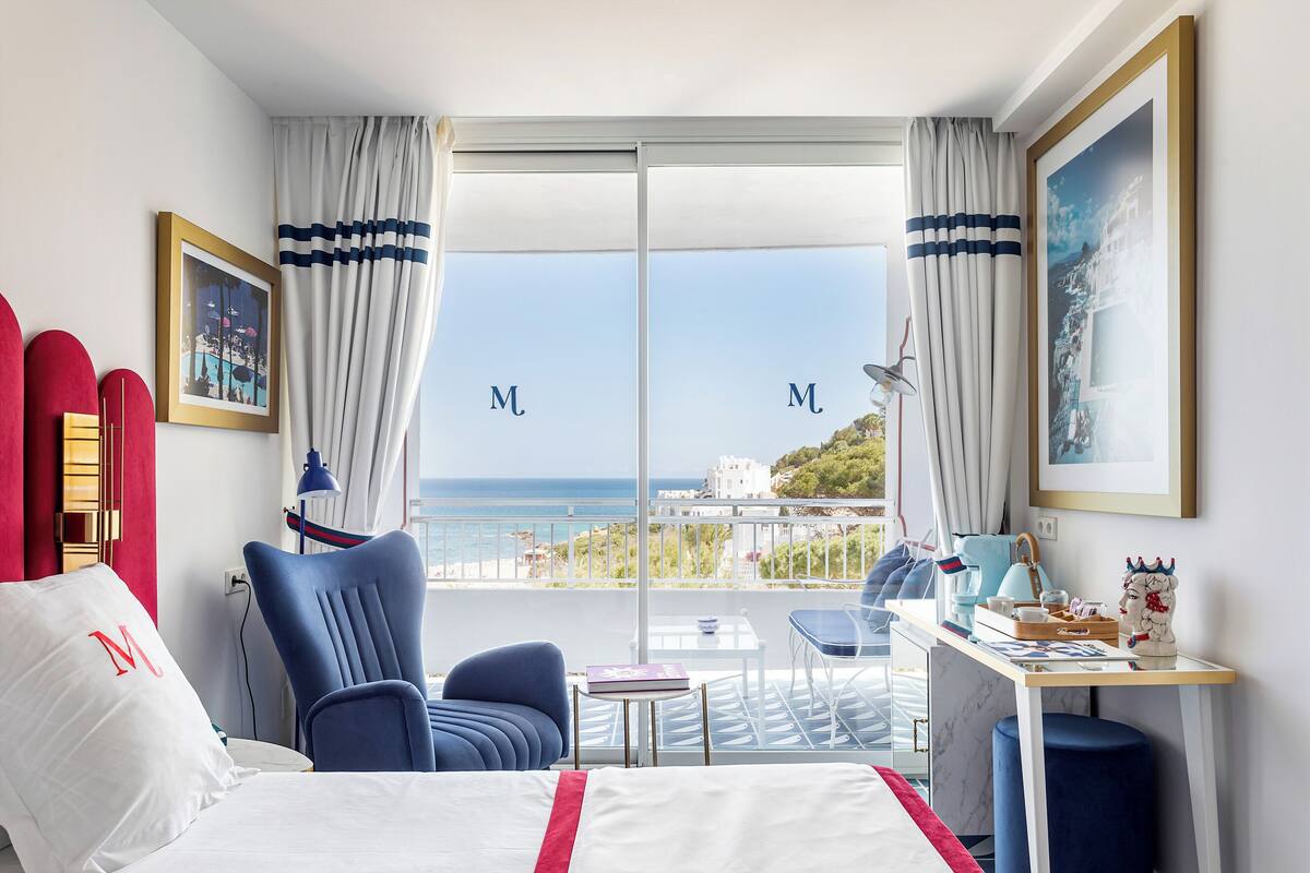 A hotel room with a bed, two chairs, a desk, and a balcony with a view of the sea.