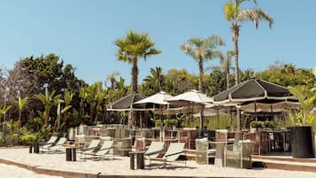 A beachside lounge with outdoor seating and umbrellas, surrounded by palm trees.