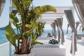 Un espace extérieur lumineux, aménagé pour accueillir des clients, avec vue sur la mer, comprenant une pergola blanche, des rideaux et des plantes tropicales.
