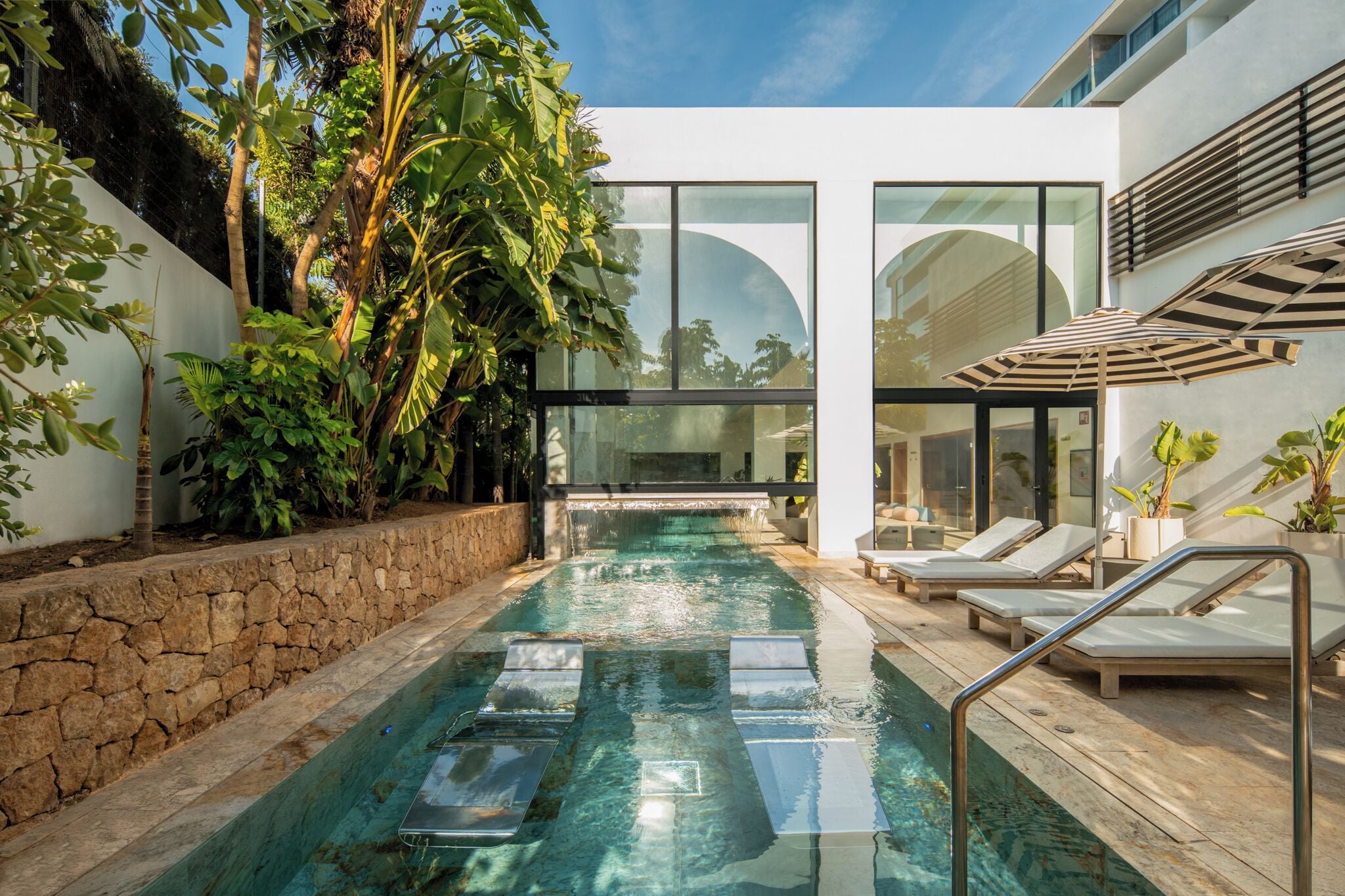 Une piscine intérieure moderne, dotée d’une eau cristalline, entourée d’une végétation tropicale luxuriante et d’un mur de pierre.