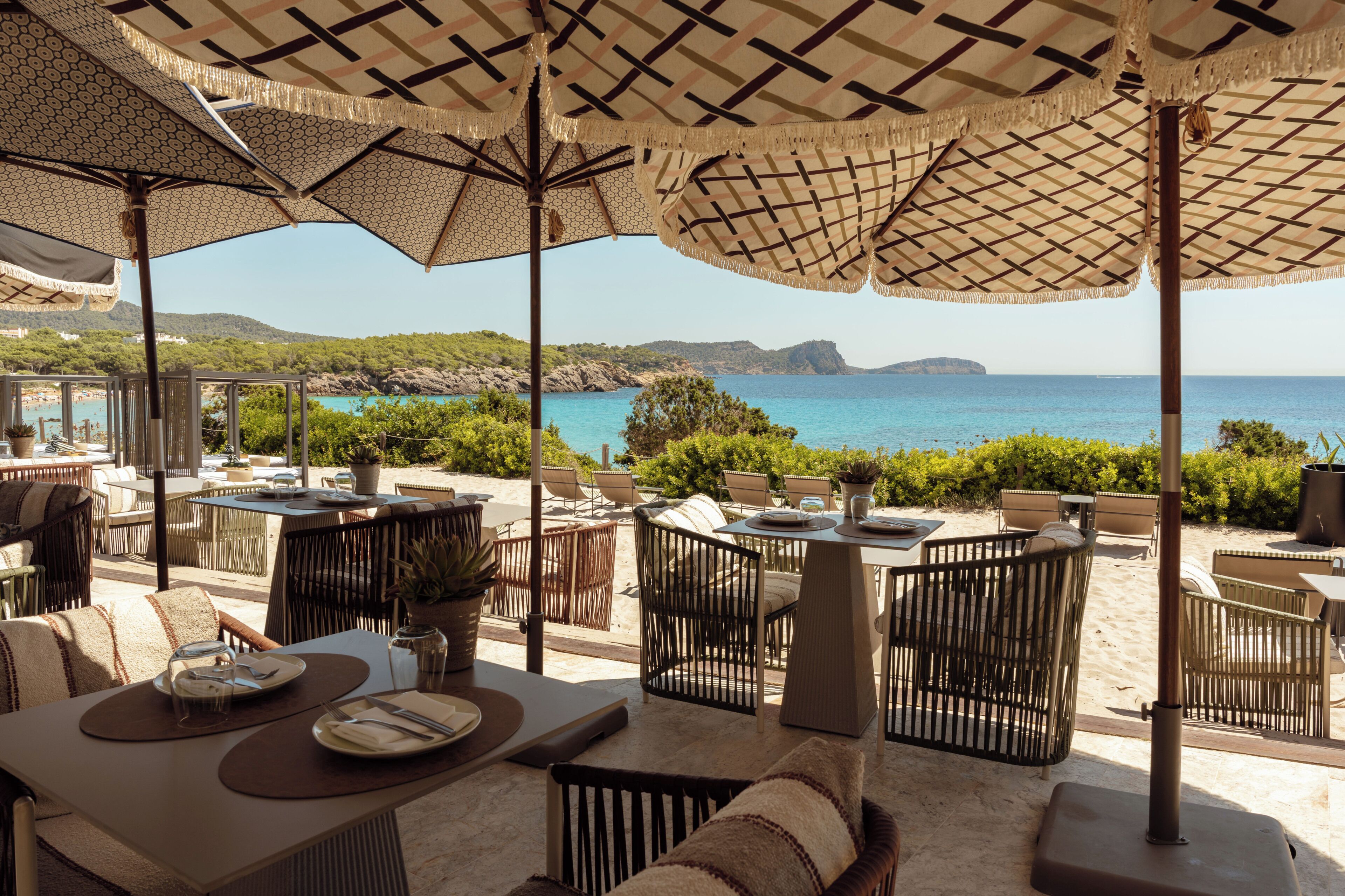 Un área de comedor al aire libre con mesas y sillas, con vistas panorámicas al mar y las colinas.