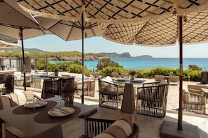 Un espace extérieur aménagé pour les repas, avec des tables et des chaises, offrant une vue panoramique sur la mer et les collines.
