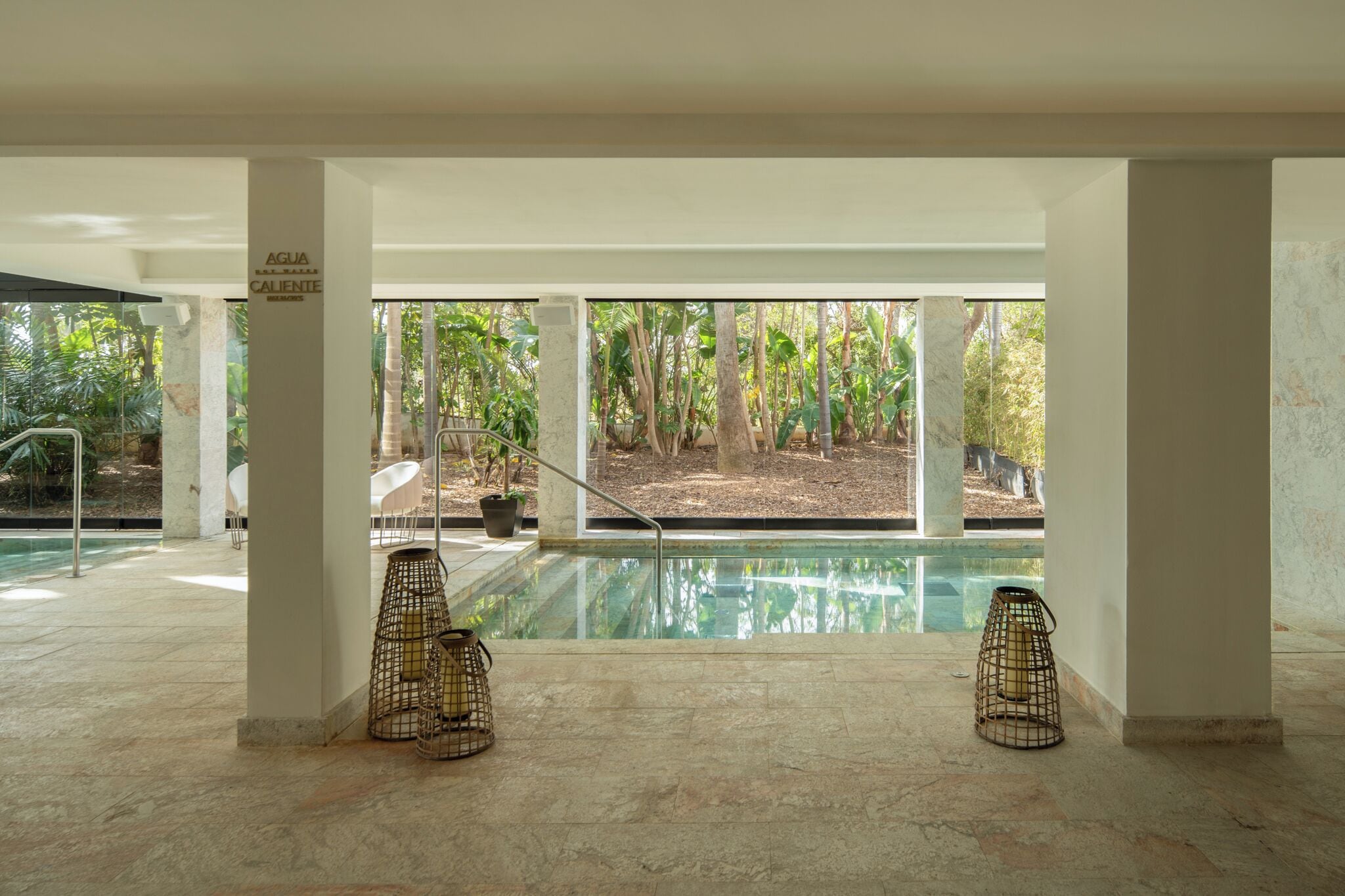 A pool area with clear views of a tropical garden, two wicker chairs, and a sign reading 'AGUA CALIENTE'.