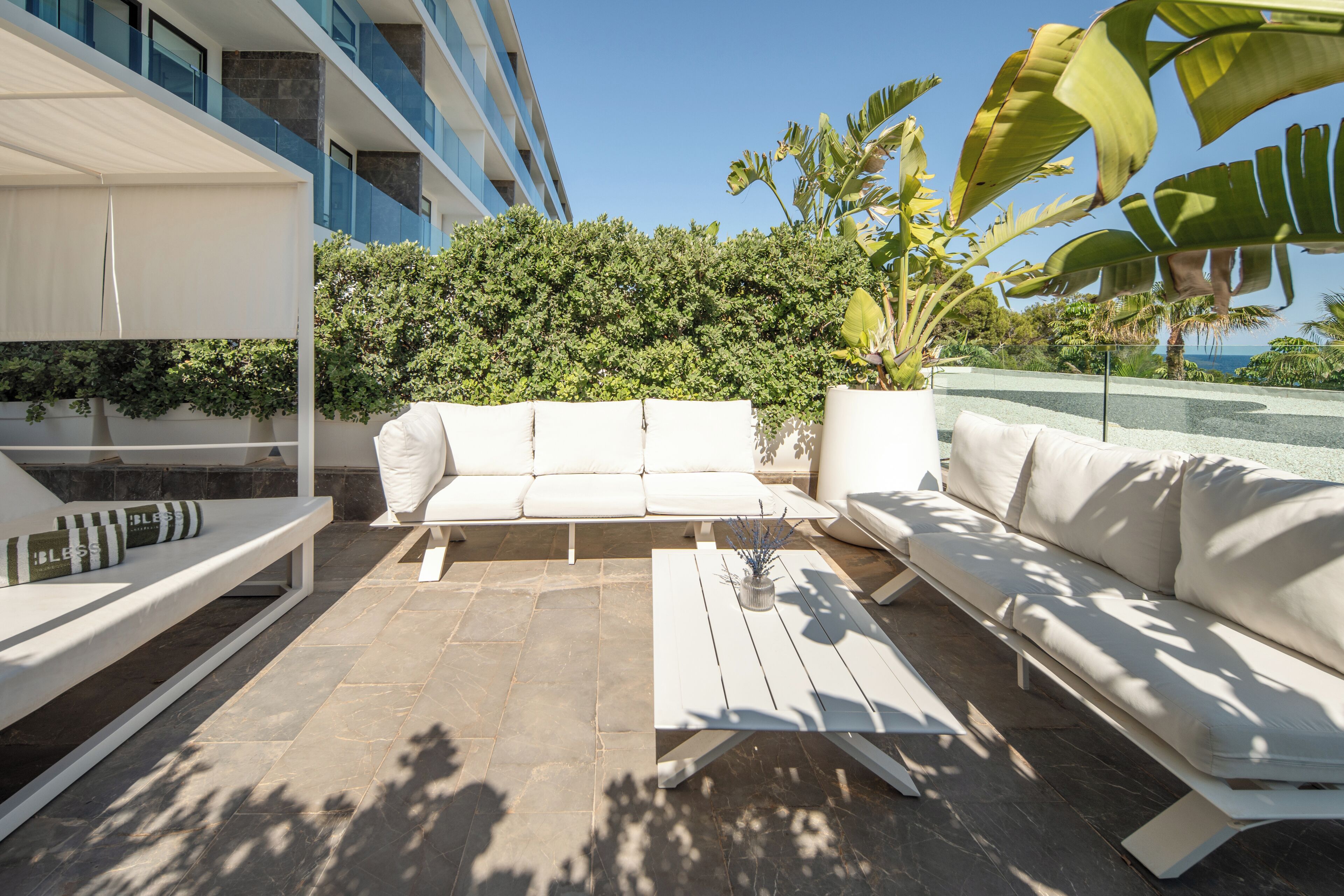 A modern outdoor seating area with white sofas and a coffee table, surrounded by a stone floor and lush greenery.