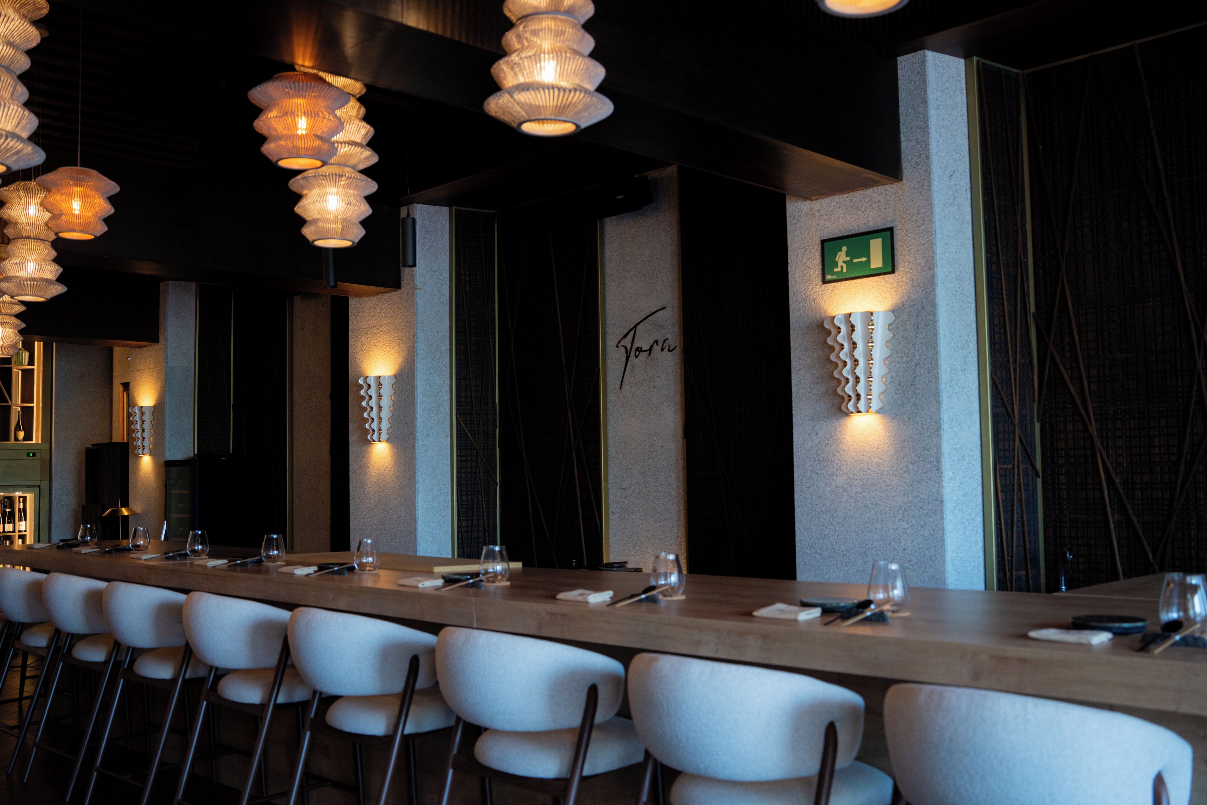 A long dining table with chairs, overhead lighting, and a wall with a sign.