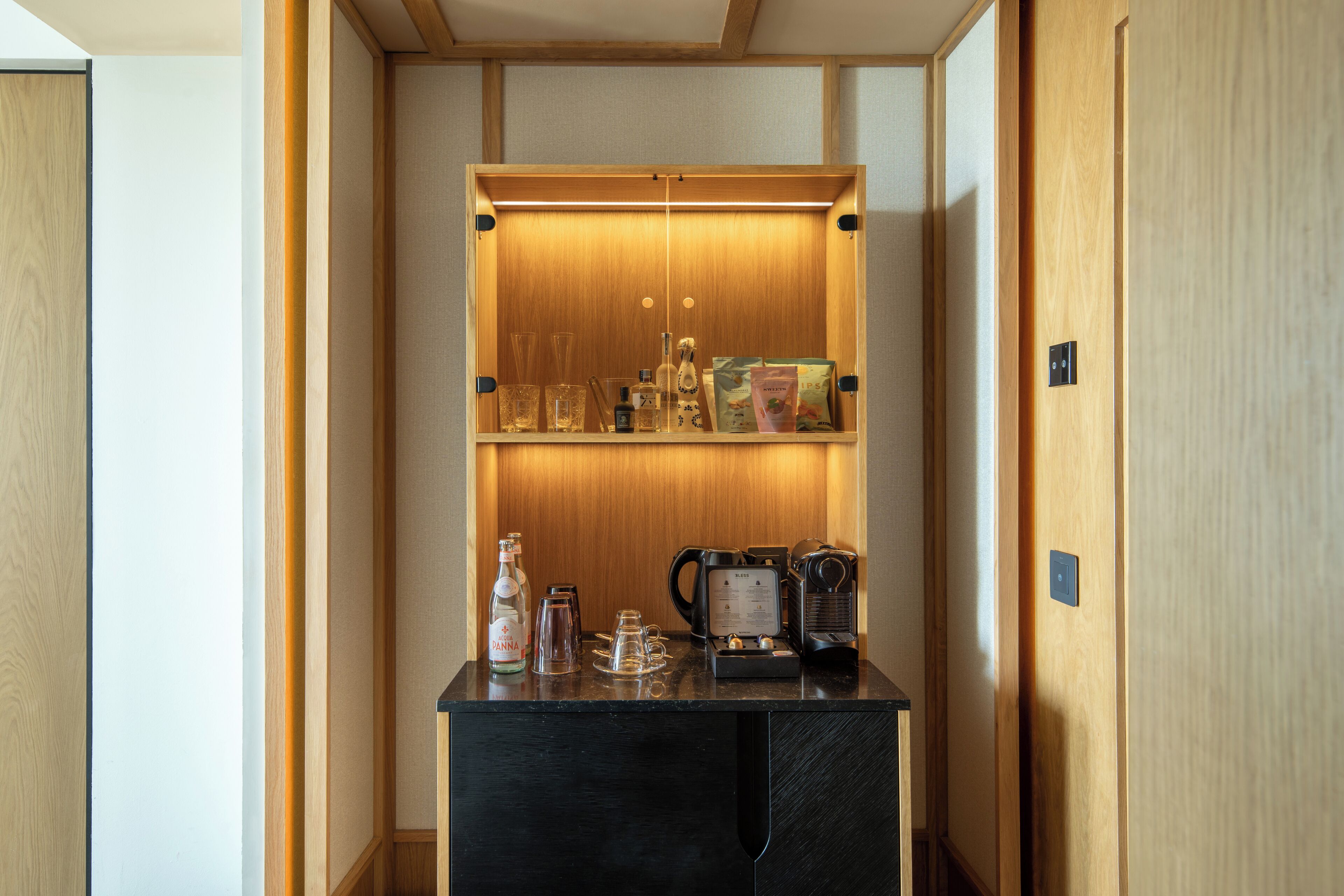 Un minibar pequeño con una cafetera, vasos y una botella.