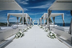 Une allée extérieure, de couleur blanche, aménagée pour un mariage, agrémentée de décorations florales et offrant une vue dégagée sur la mer.