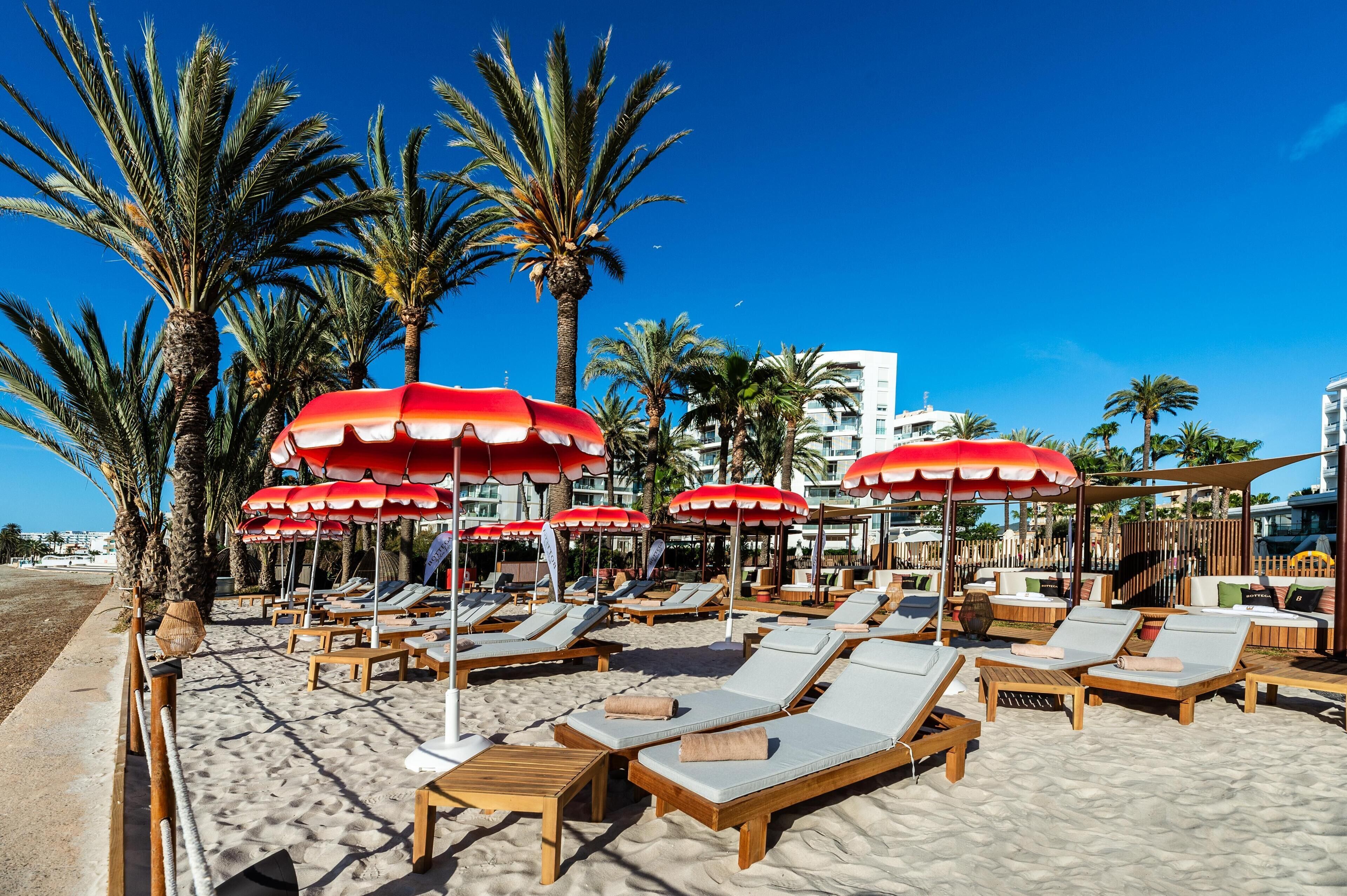 A beachside lounge area with sun loungers and red umbrellas, palm trees, and a clear blue sky.