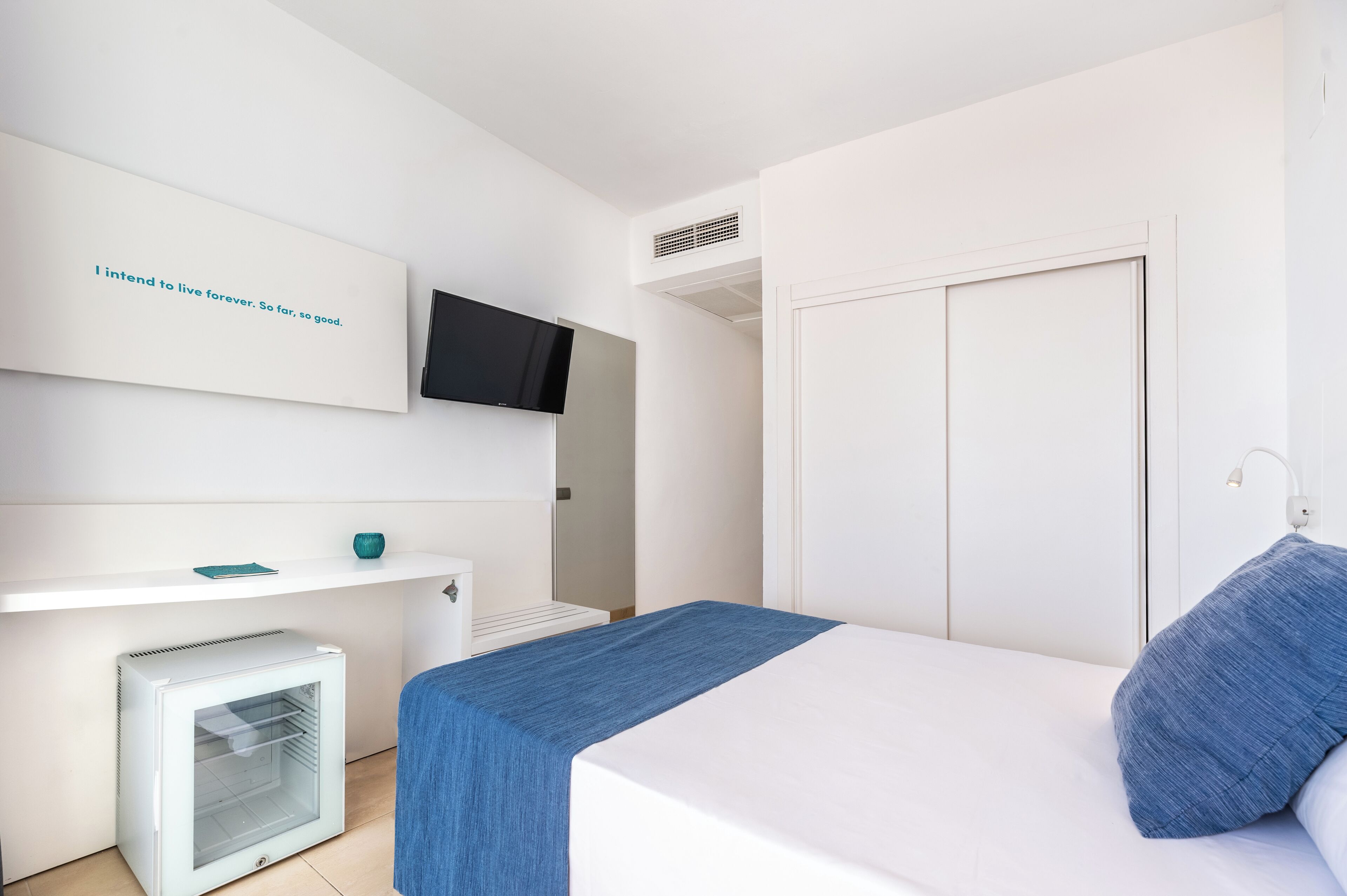 Un dormitorio moderno con una cama, un televisor montado en la pared, un refrigerador pequeño y un armario blanco.