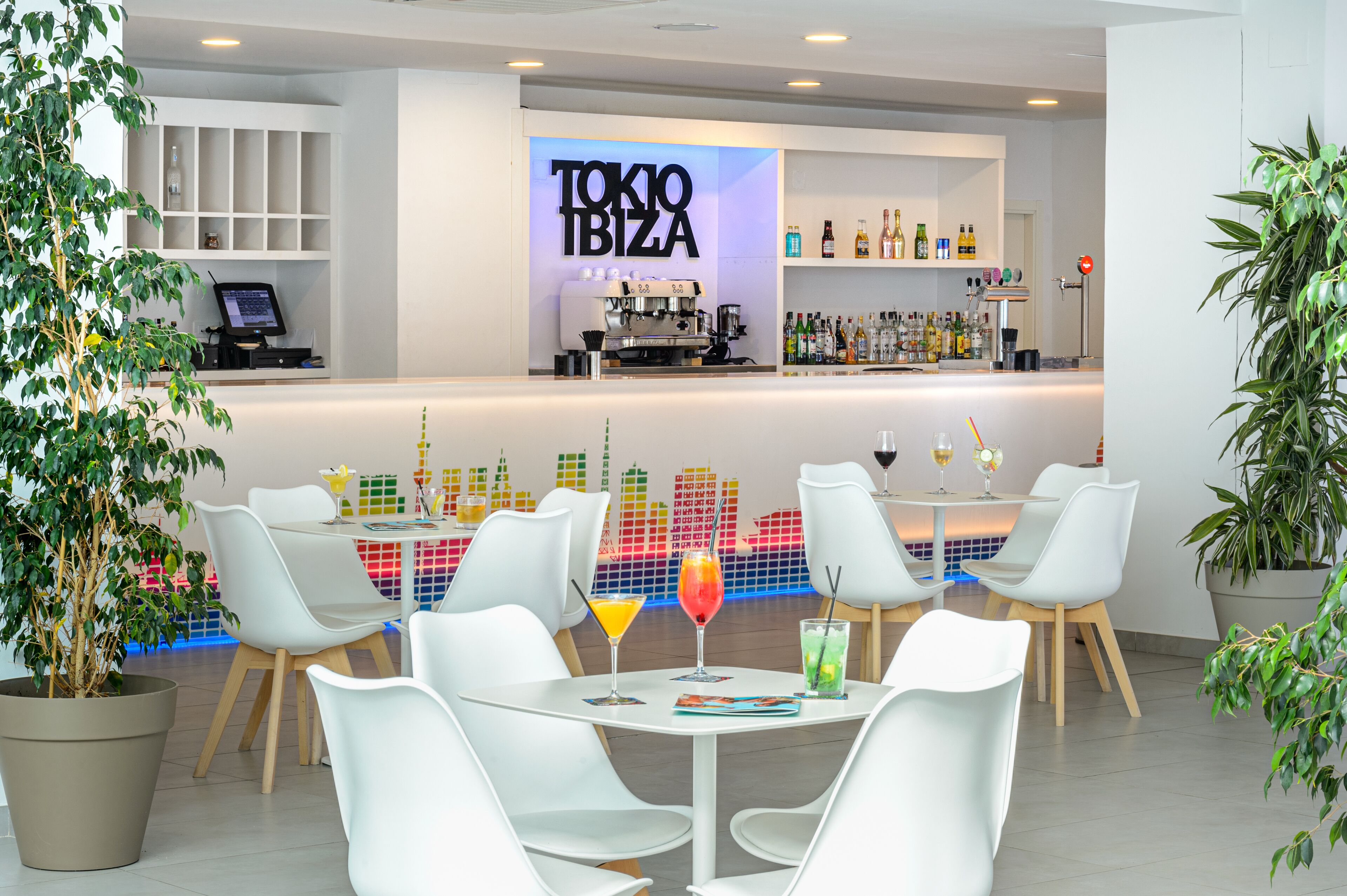 A modern bar area with a neon sign reading "TOKYO IBIZA," white chairs, and a variety of drinks on the tables.