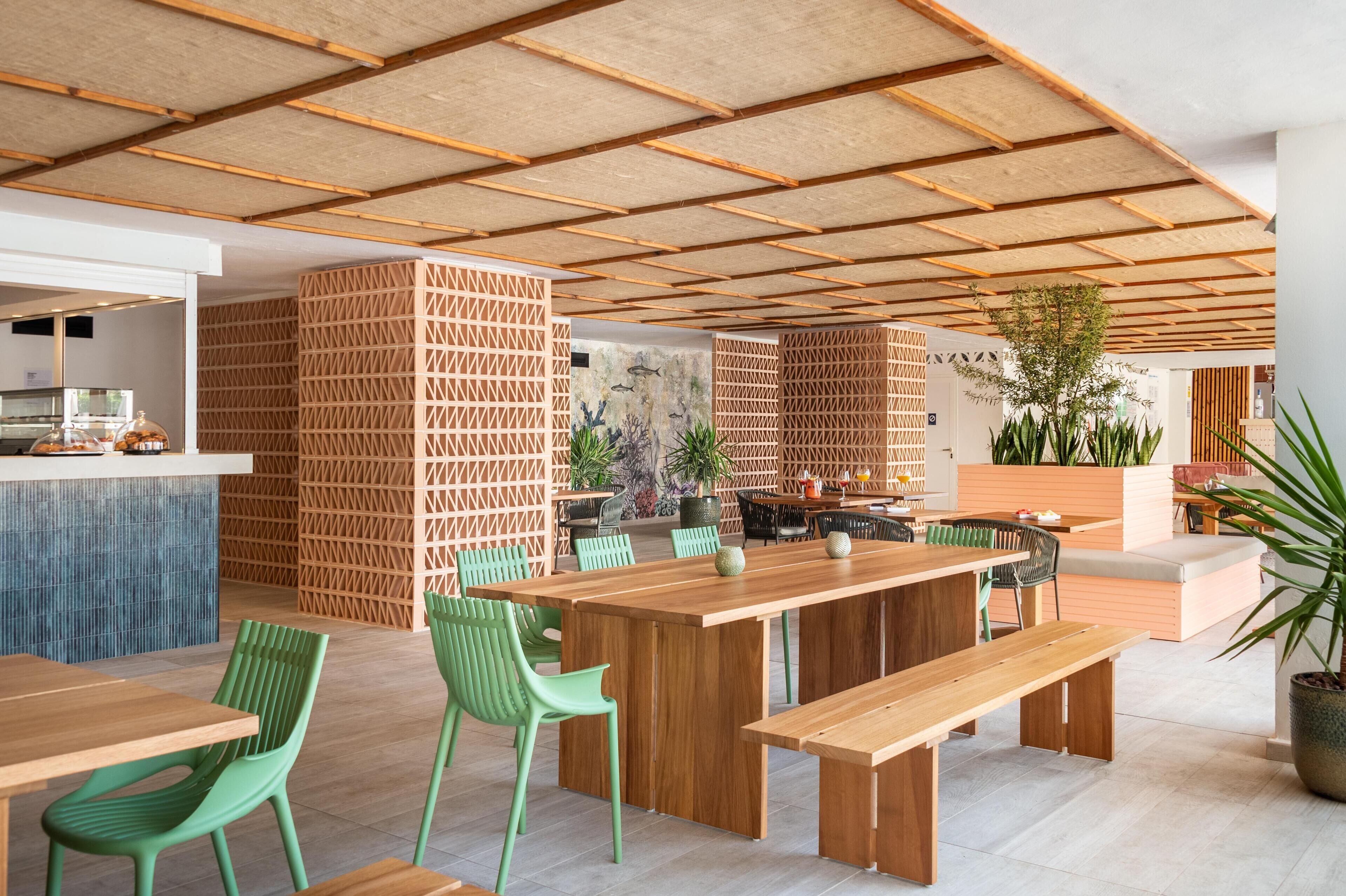 Un restaurante moderno con mesas de madera y sillas verdes, una zona de cocina y un área de estar con plantas.