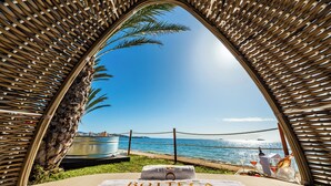 A beach view through a woven structure with a towel labeled "Bottega" and a table with drinks and a bottle. - Hotel Vibra Algarb (Ibiza Town)