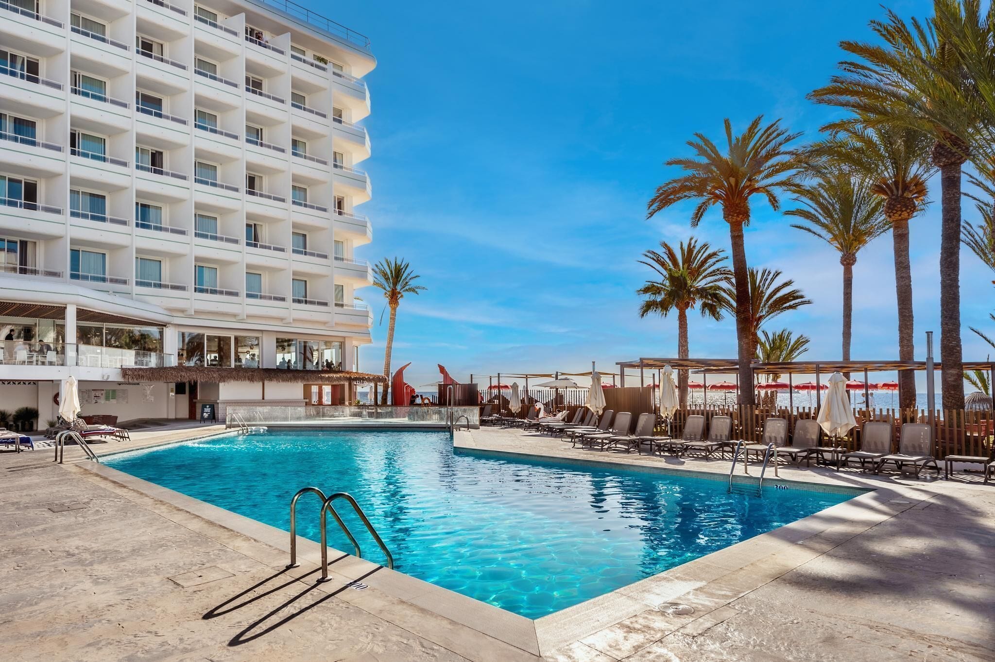 A hotel pool area with a clear blue pool, lounge chairs, and palm trees.