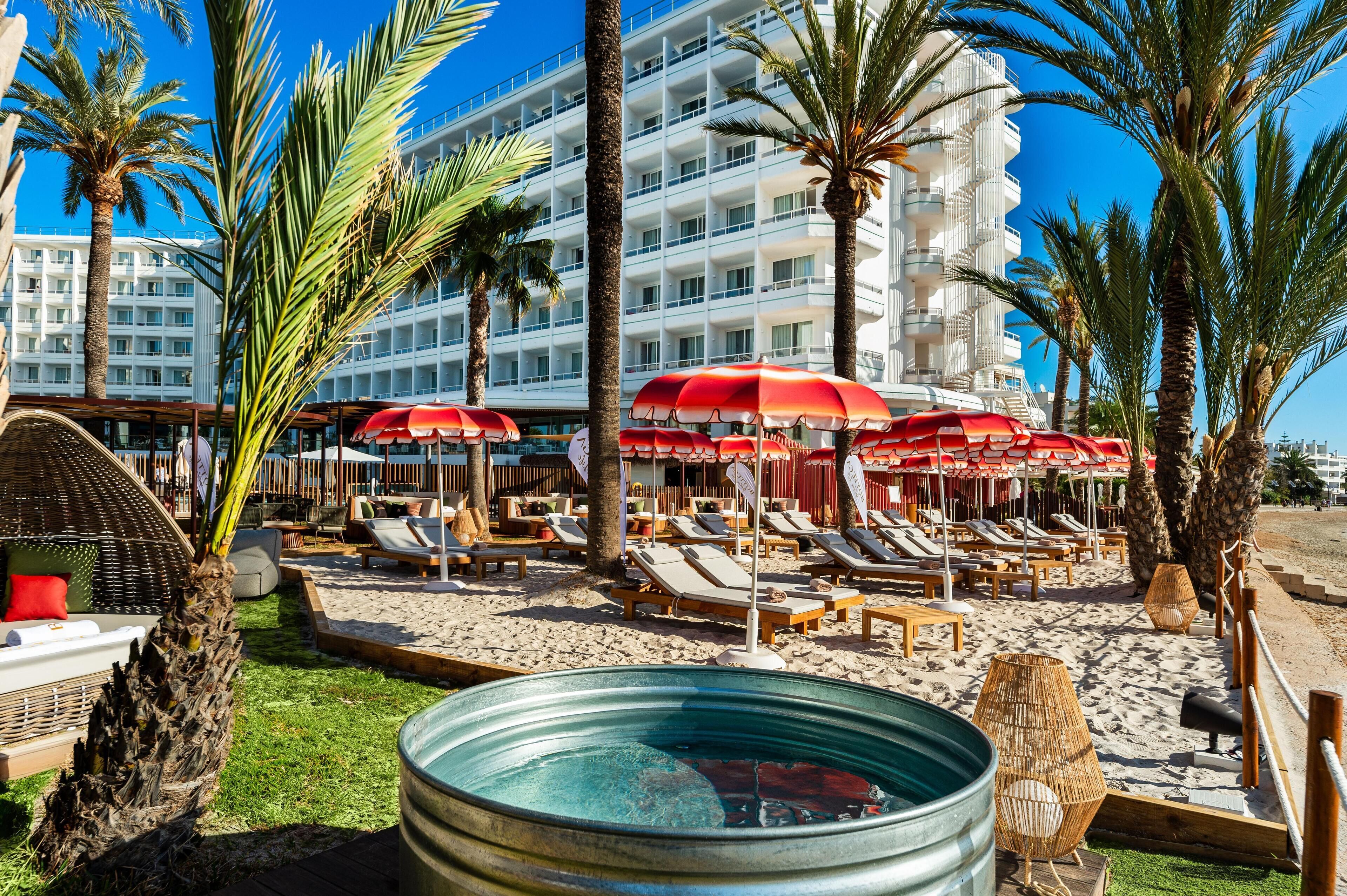Un hôtel situé en bord de mer, doté de chaises longues, de parasols, d’une piscine et de palmiers.