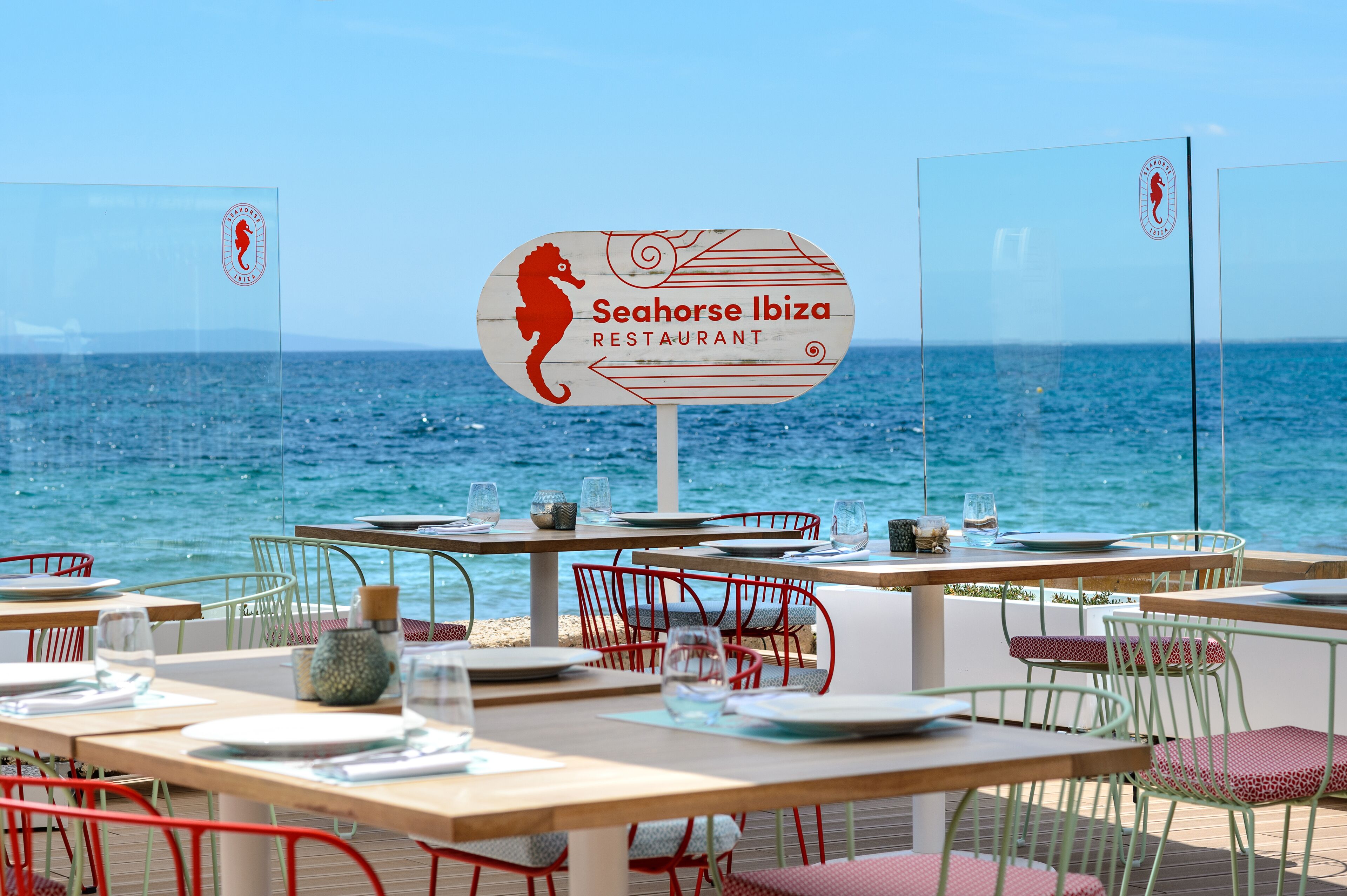 Um restaurante à beira-mar com um letreiro que diz 'Seahorse Ibiza Restaurant', mesas com cadeiras e vista para o mar.